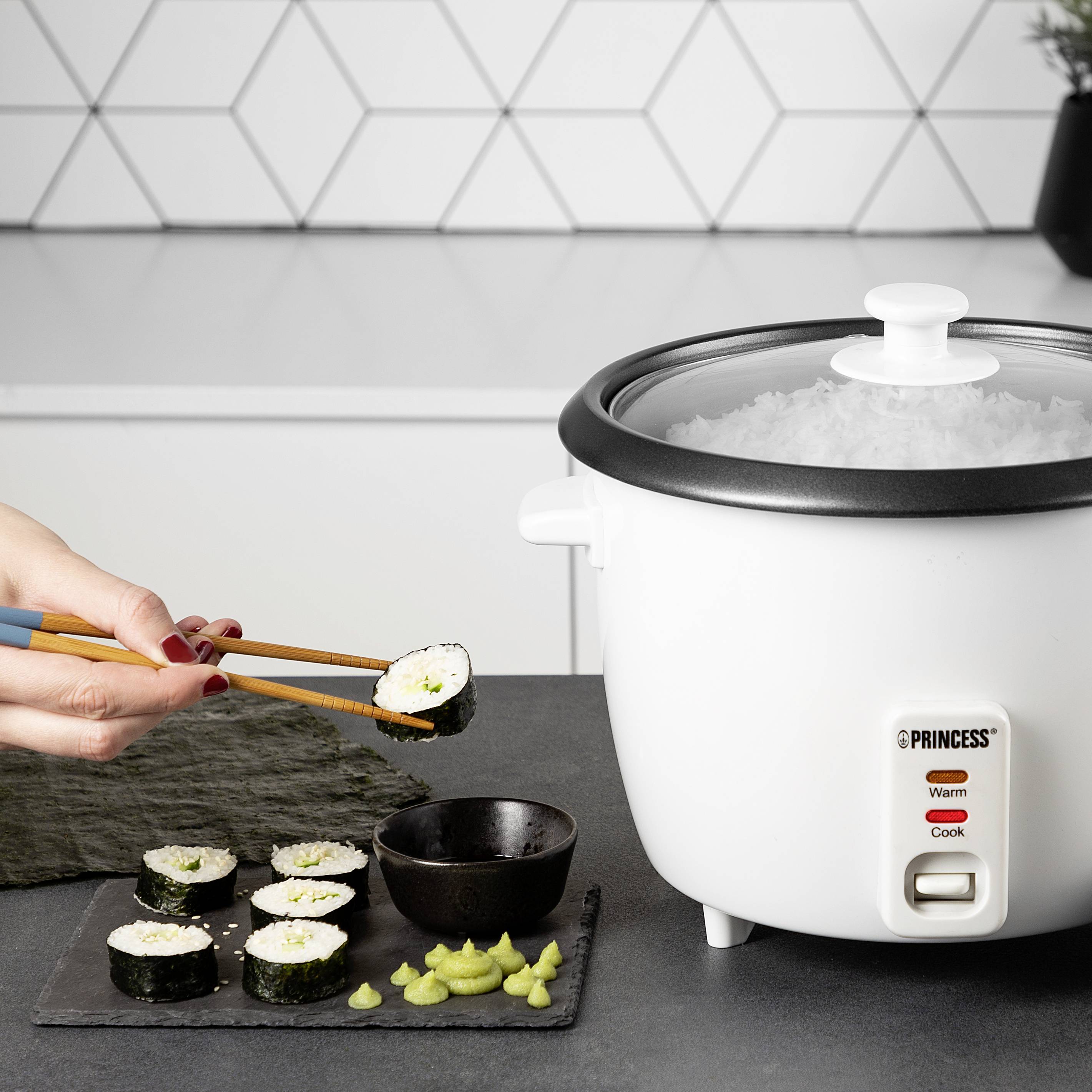 Osoba drží sushi rolky pomocí jídelních hůlek nad stolem s rýžovarem, wasabi a sójovou omáčkou. V pozadí bílé obklady.