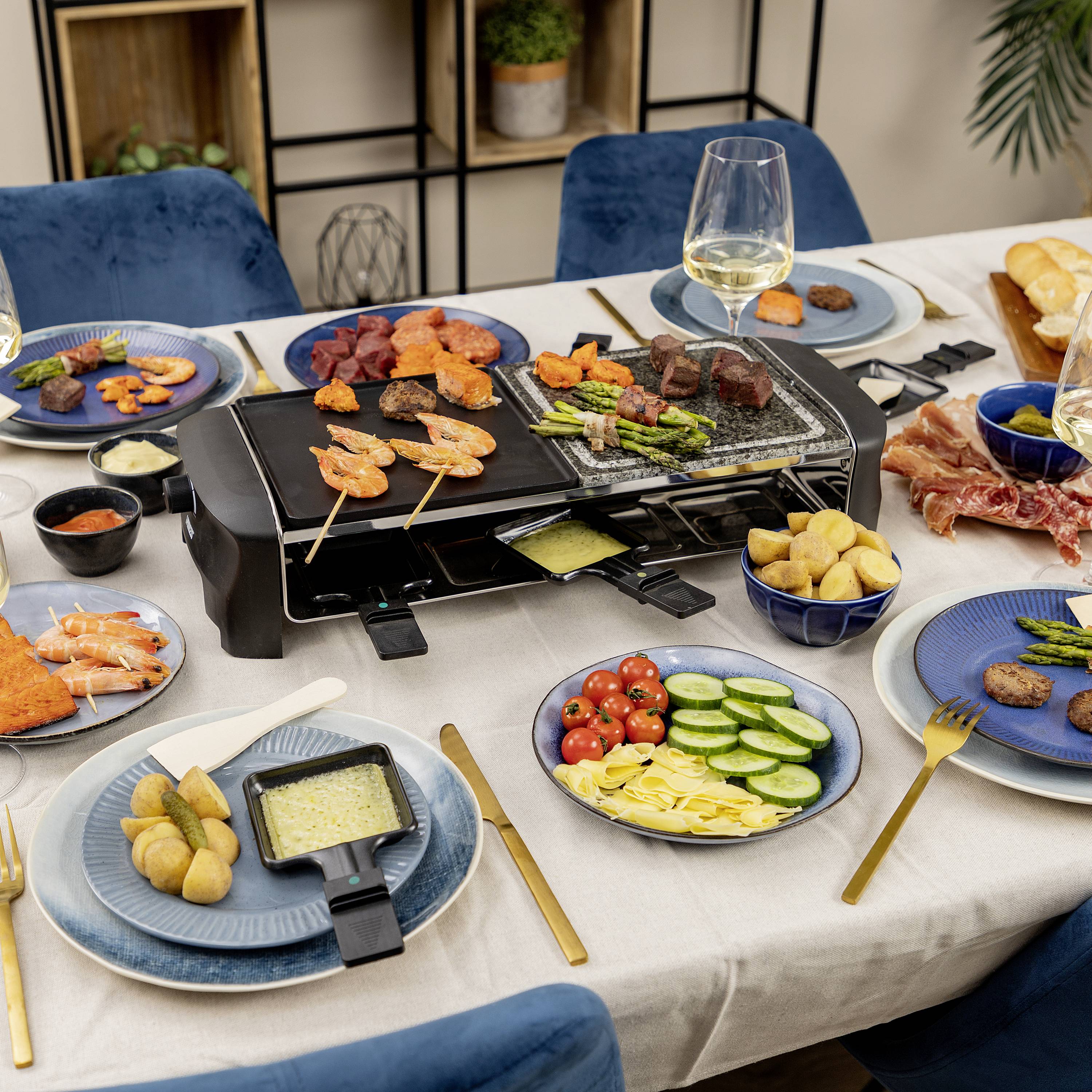Prostřený stůl s raclette grilem, na kterém se opékají zelenina a maso. Talíře s přílohami jako je chléb, salám a zelenina jsou rozmístěné.