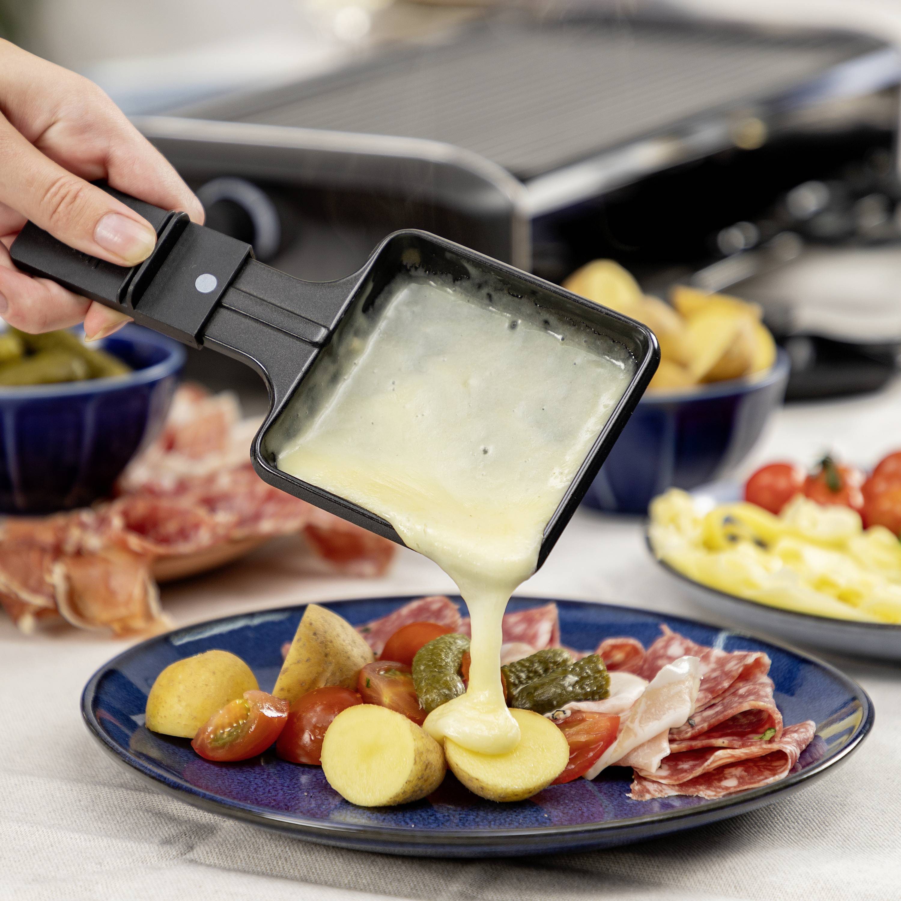 Osoba nalévá roztavený sýr na raclette s bramborami, nakládanými okurkami a uzeninou. V pozadí je vidět gril.