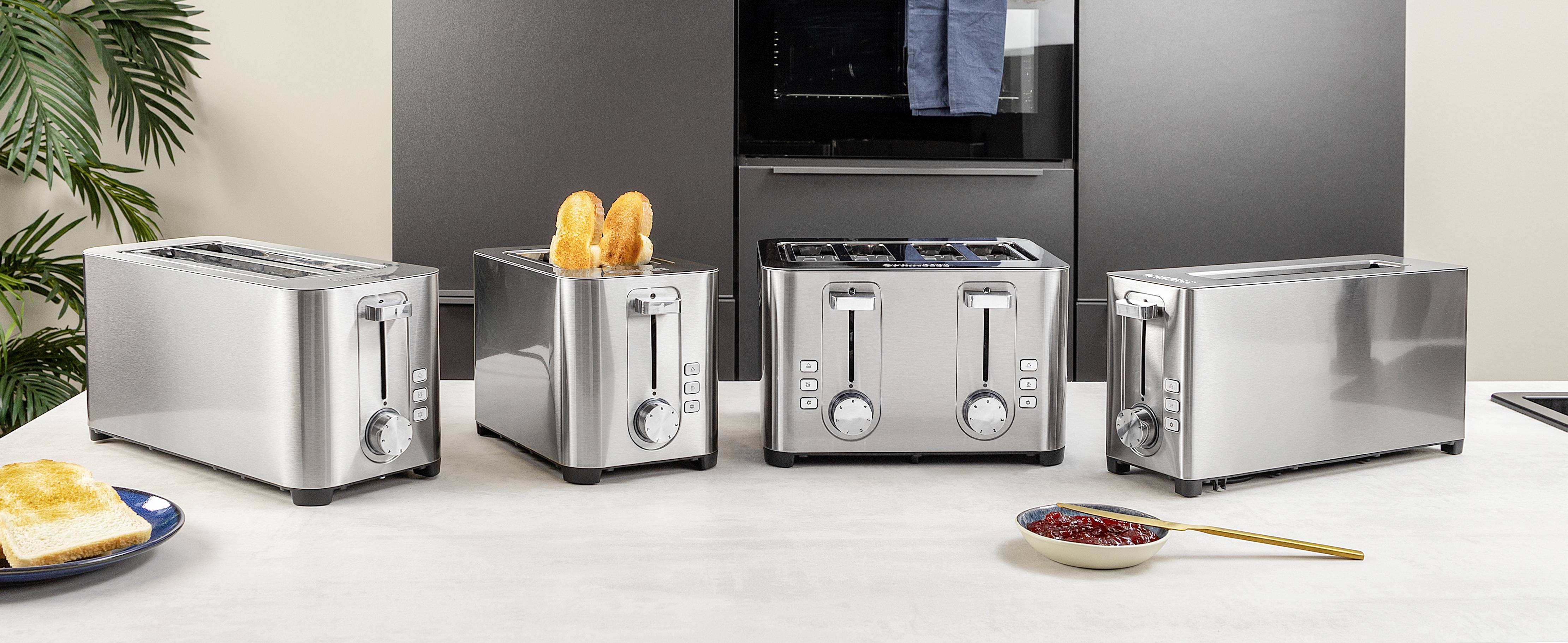 Čtyři stříbrné toustovače na kuchyňské lince, jeden s kousky chleba. V pozadí černá kuchyňská skříňková stěna.