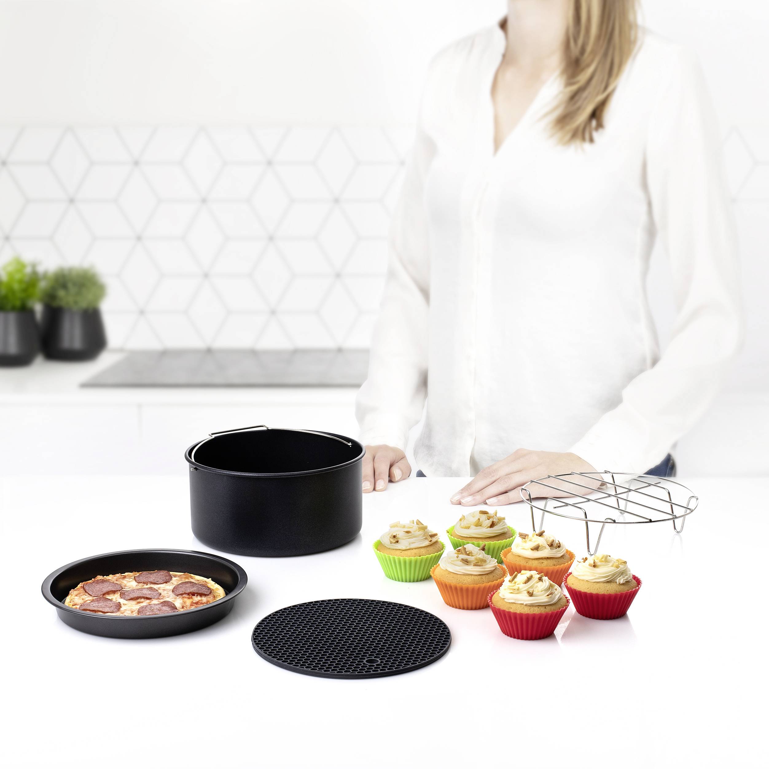 Osoba v bílém horním dílu oblečení stojí u stolu s pizzou, barevnými muffiny, černou pánví a podtácky.