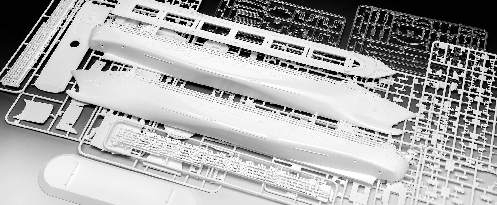 Několik bílých plastových rámečků s detailními komponenty modelářské stavby lodě, rozprostřených na tmavém pozadí.