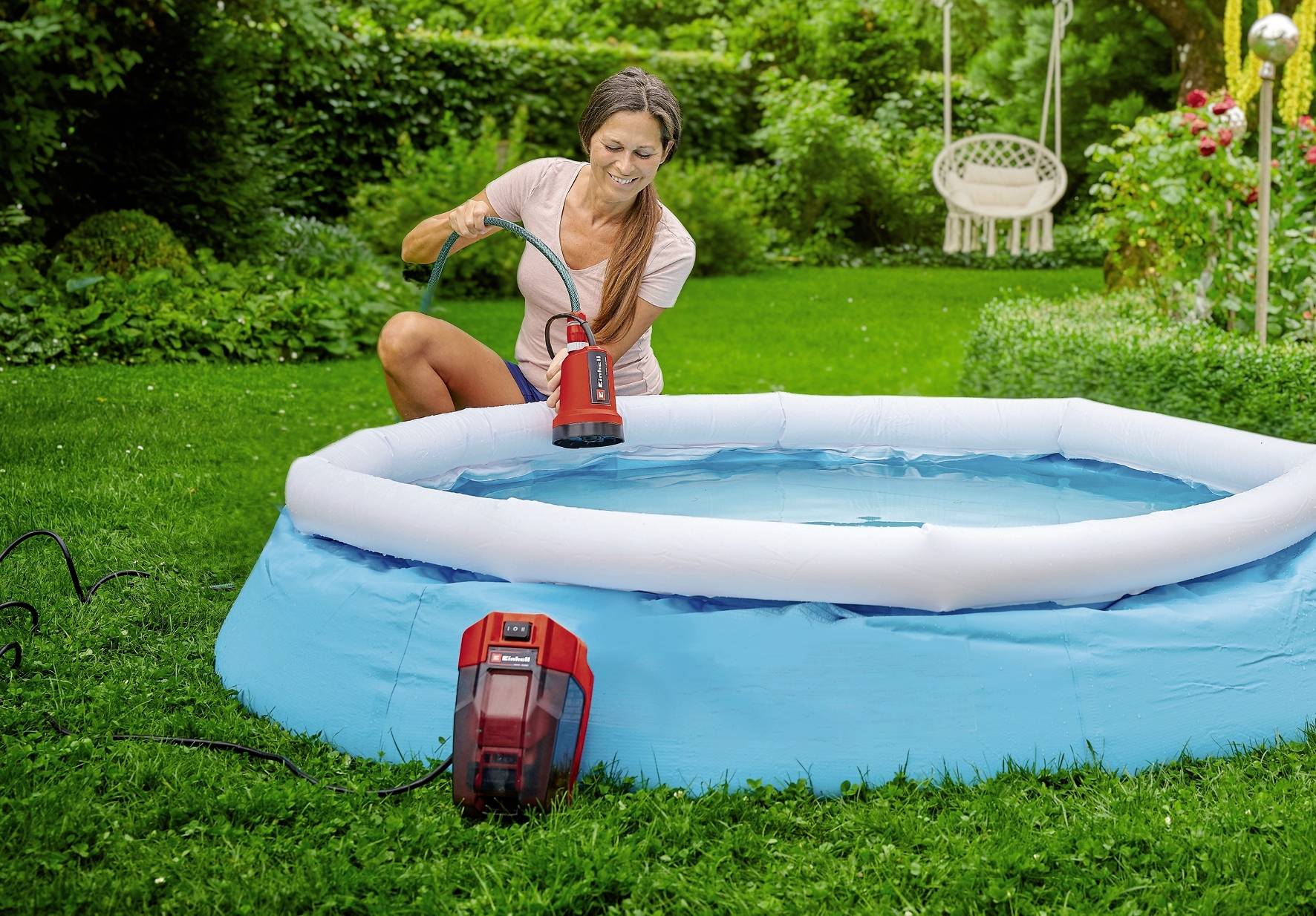 Žena čerpá elektrickým čerpadlem vodu do nafukovacího bazénu na zahradě. V pozadí je vidět hodně zeleně.