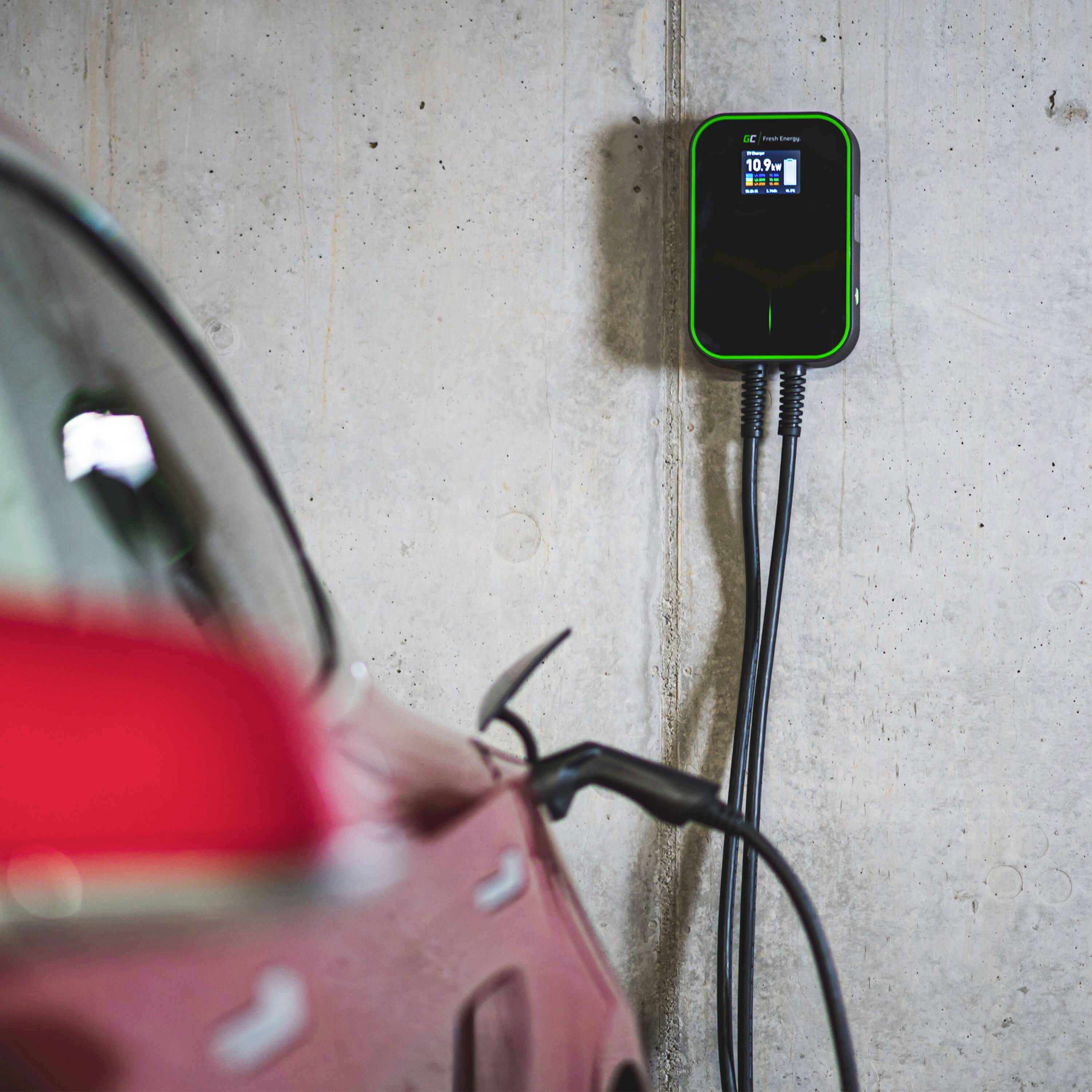 Červené elektroauto je připojeno k nabíjecí stanici namontované na stěně, displej na stanici zobrazuje stav baterie.