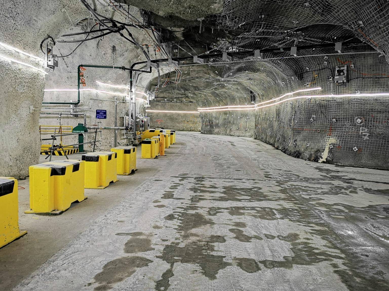 Dlouhý podzemní tunel s osvětlenými stropy a žlutými bariérami po stranách. Podlaha je betonová a vlhká.