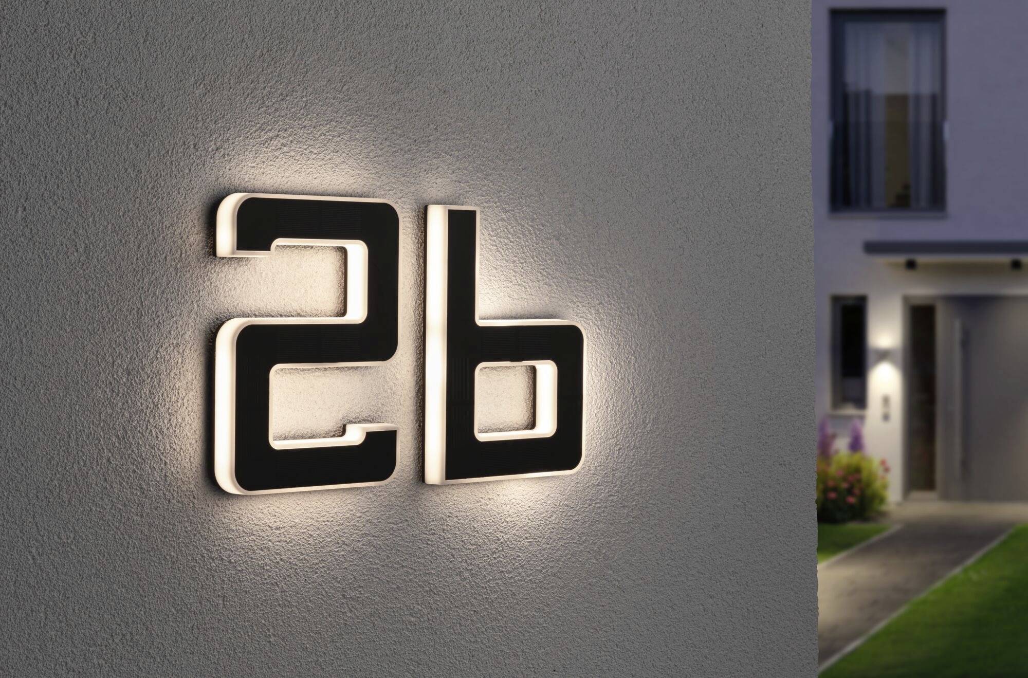 Domovní číslo '26b' na zdi, jemně osvětlené. V pozadí vchod a osvětlená cesta vedoucí k budově.