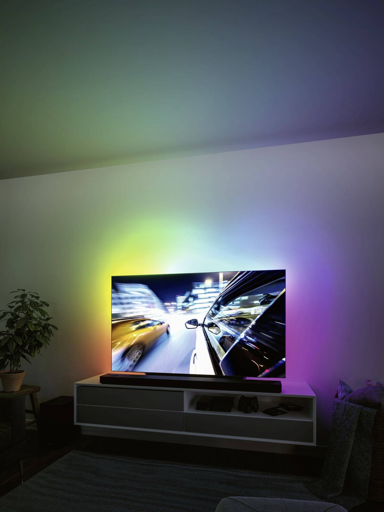 Televizor zobrazuje rozmazaný obraz jedoucího automobilu v noci, s barevnými světly osvětlujícími stěnu.