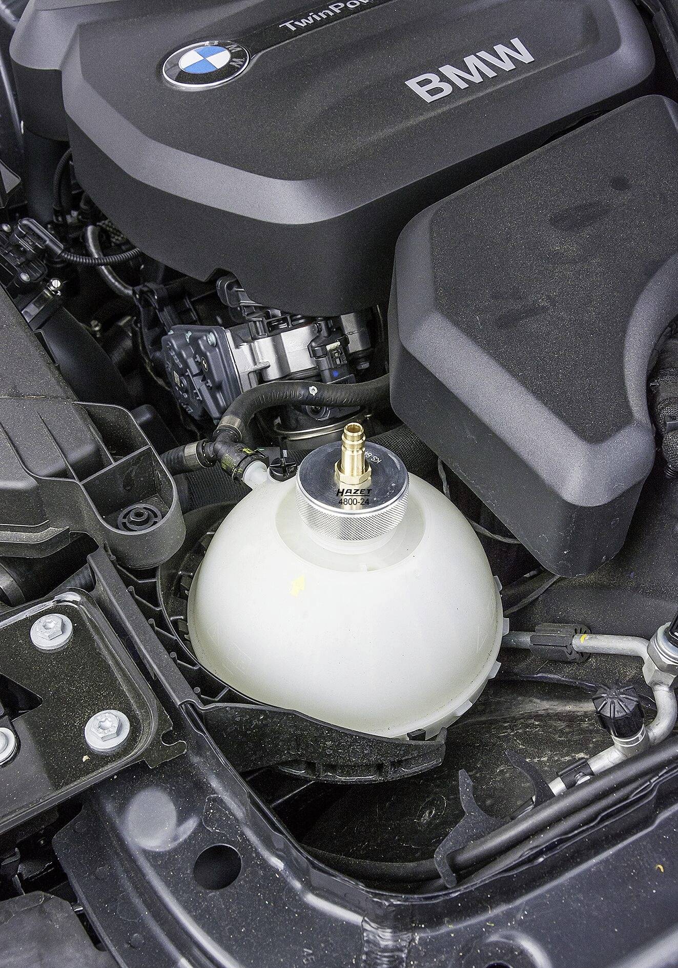 Bílá nádobka na kapalinu v motorovém prostoru BMW, obklopená různými komponenty motoru.