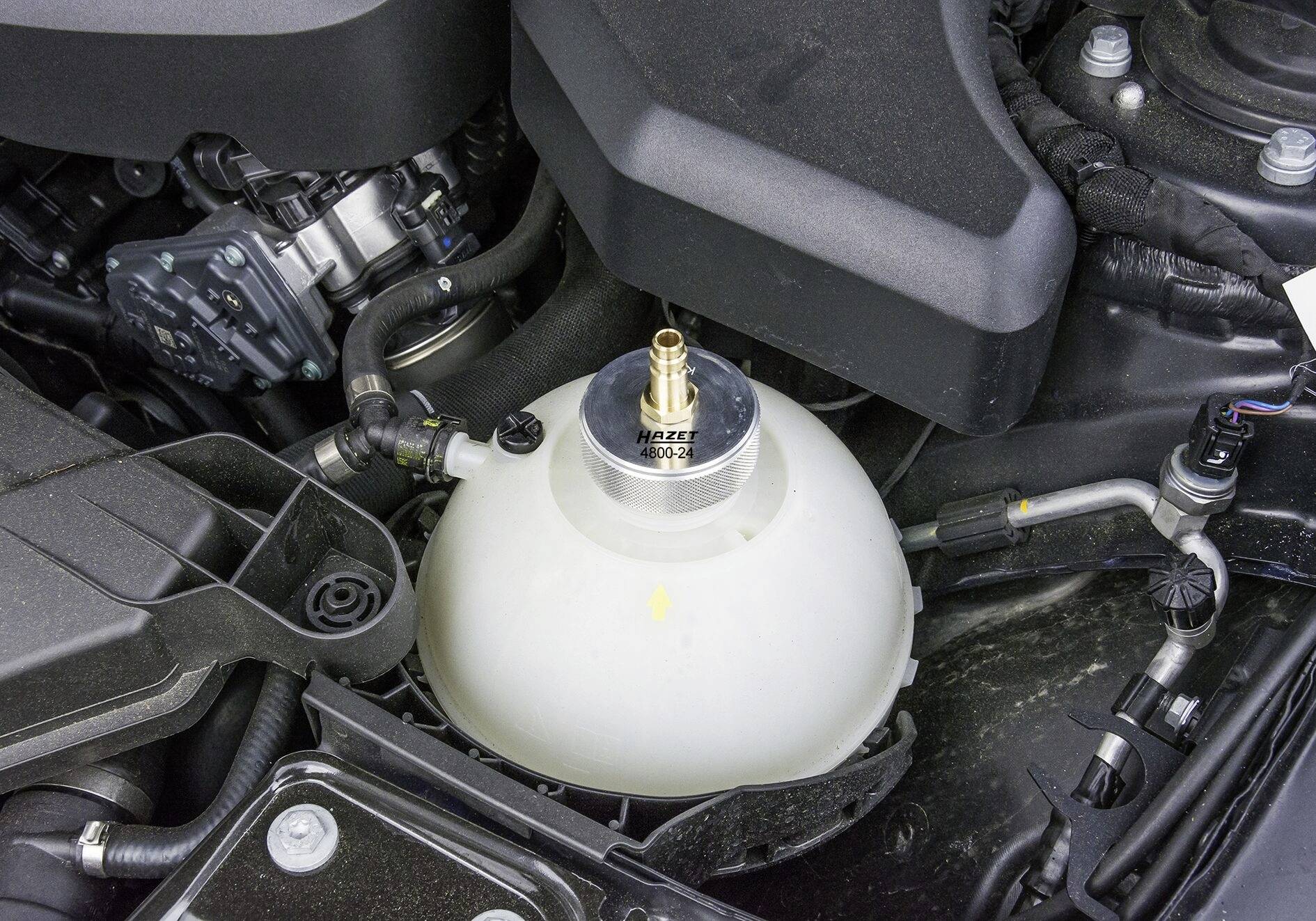 Nádobka chladicí kapaliny automobilu v motorovém prostoru, s připojenými hadicemi a viditelnými komponenty motoru v pozadí.