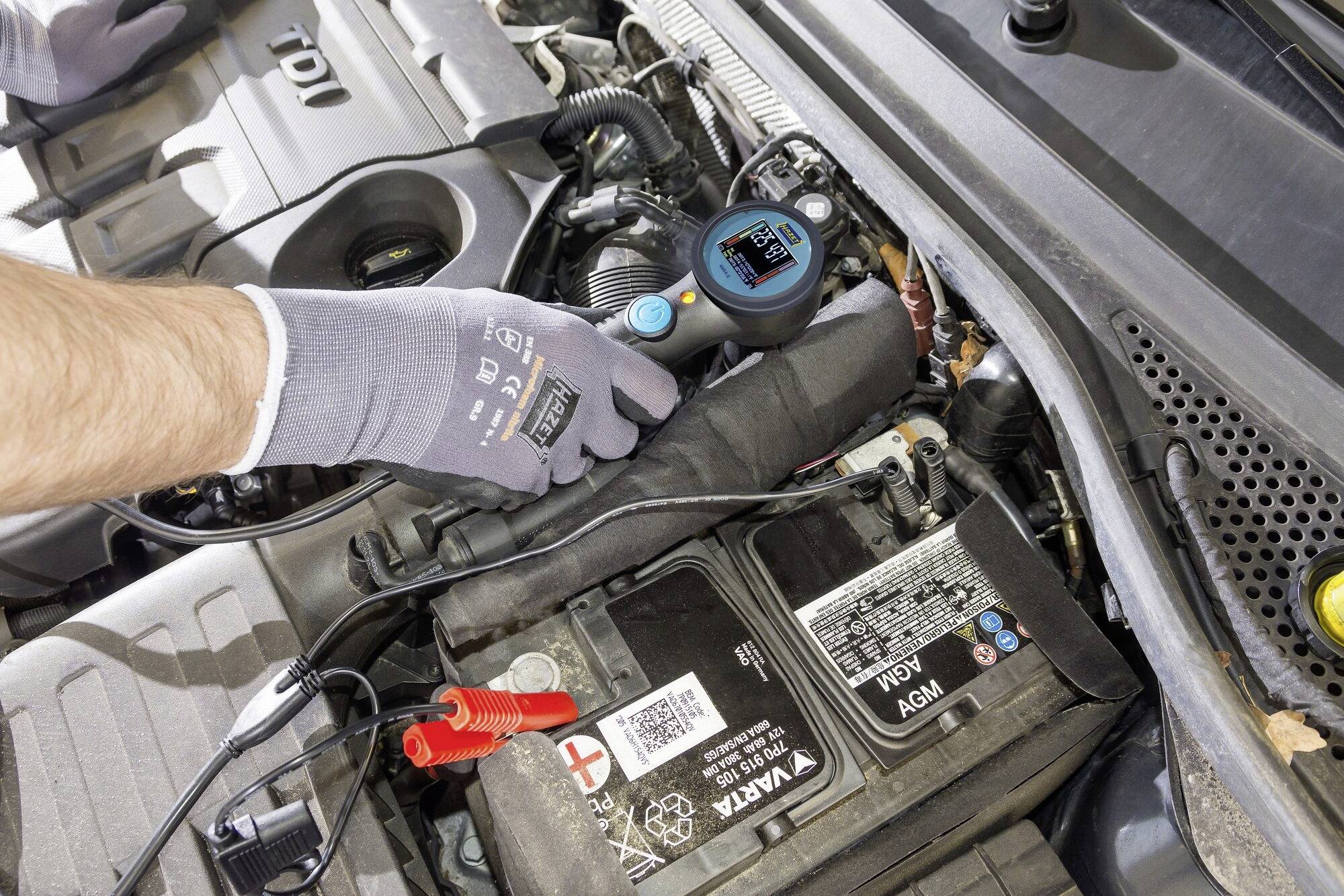 Osoba kontroluje baterii automobilu diagnostickým zařízením. Přístroj ukazuje stav nabití. Motorová kapota je otevřená.