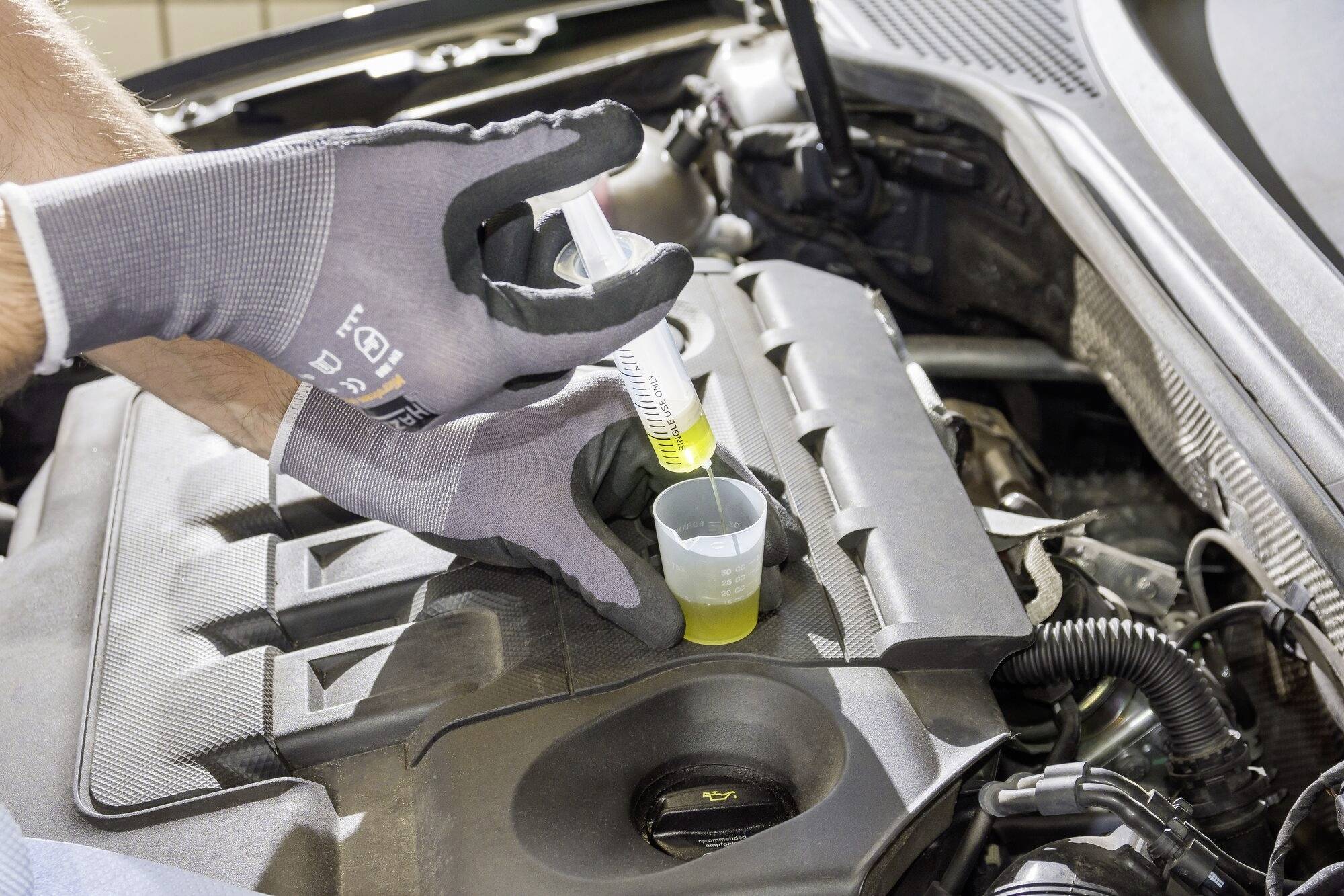 Mechanik plní žlutou kapalinu ze stříkačky do plastového kelímku v otevřené nádrži automobilu v motorovém prostoru.