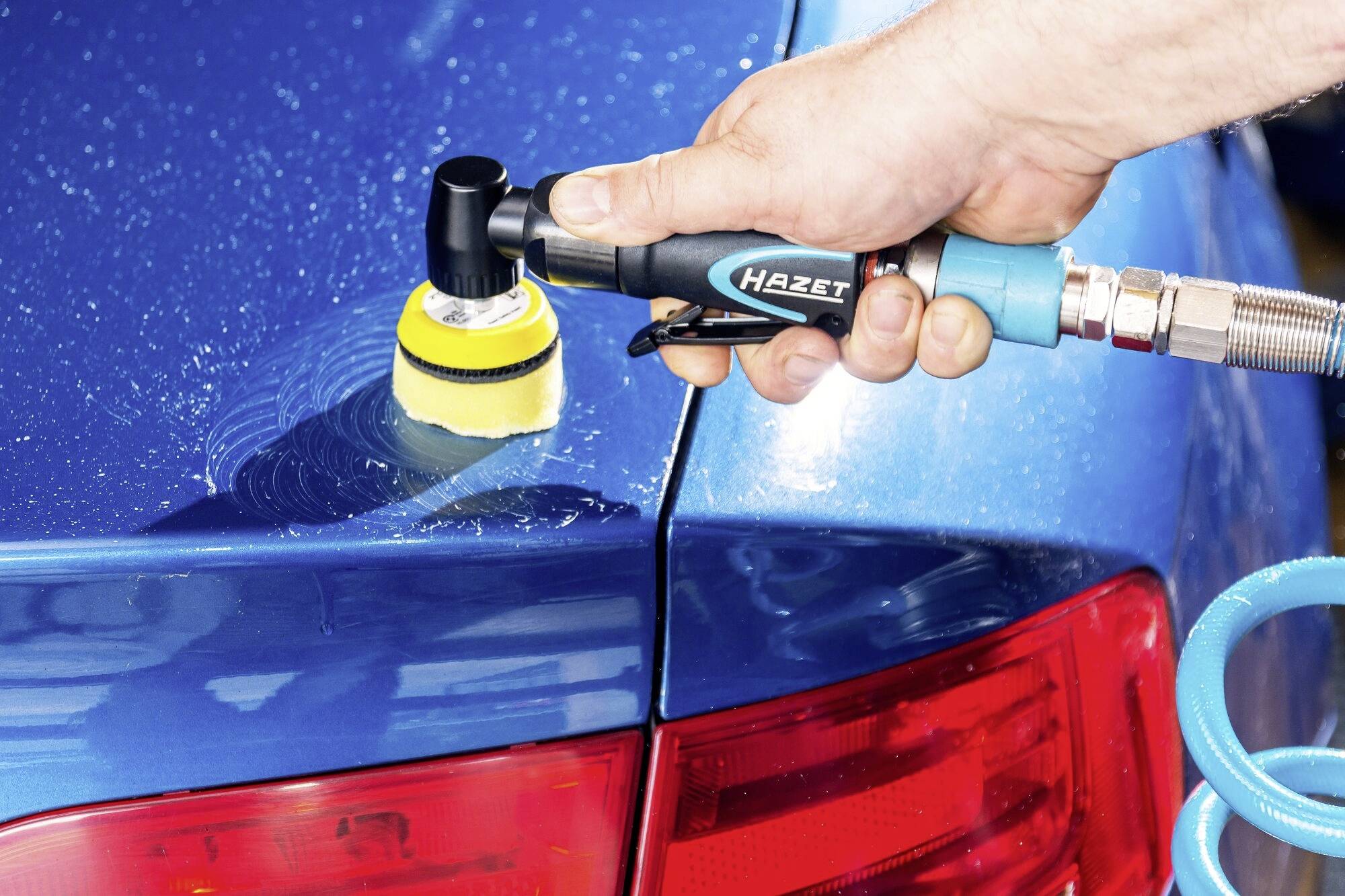 Ruka leští modré lakování automobilu elektrickým leštícím přístrojem v detailním záběru.