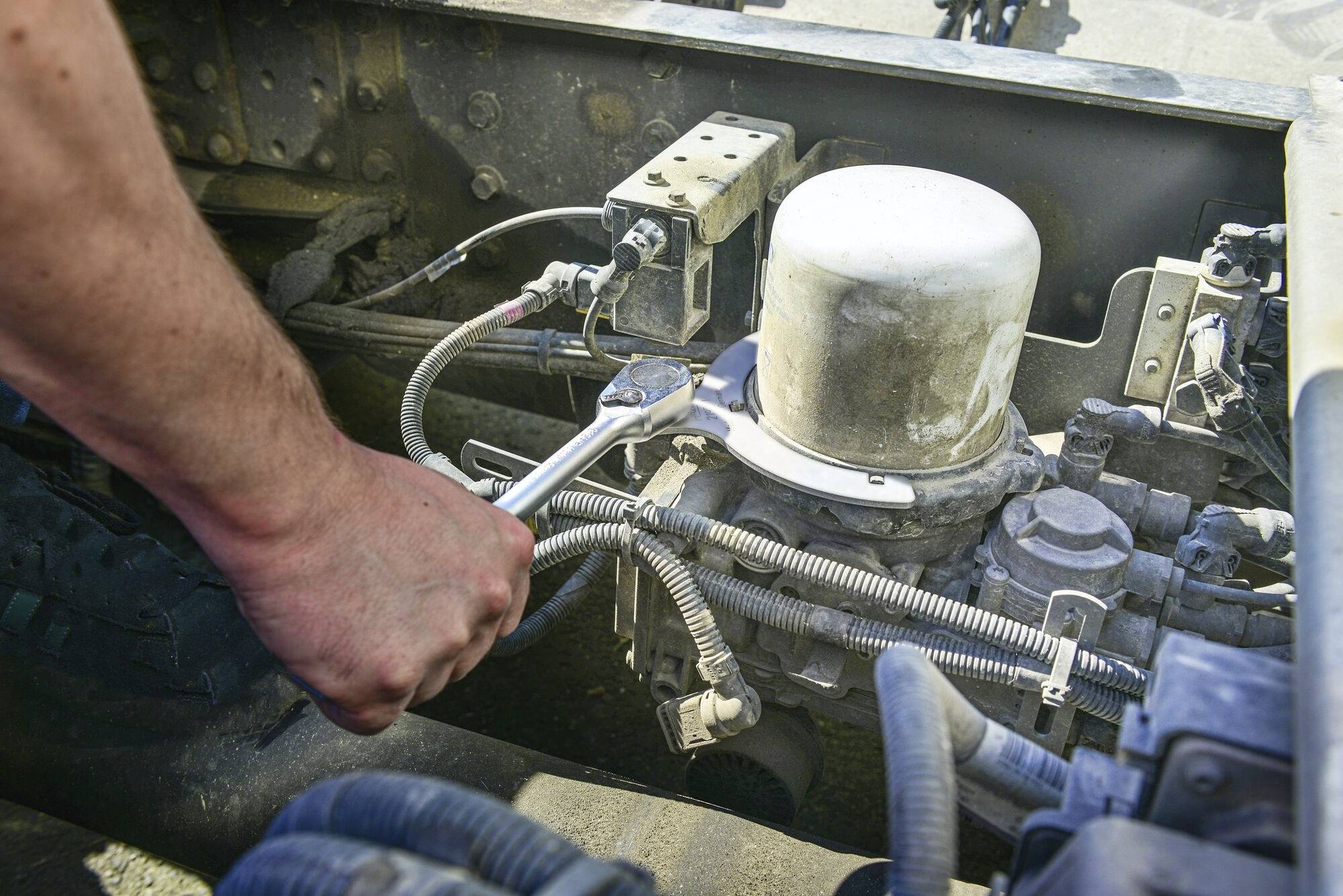 Osoba utahuje šroub pomocí klíče v motoru vozidla, obklopena kabely a mechanickými součástkami.