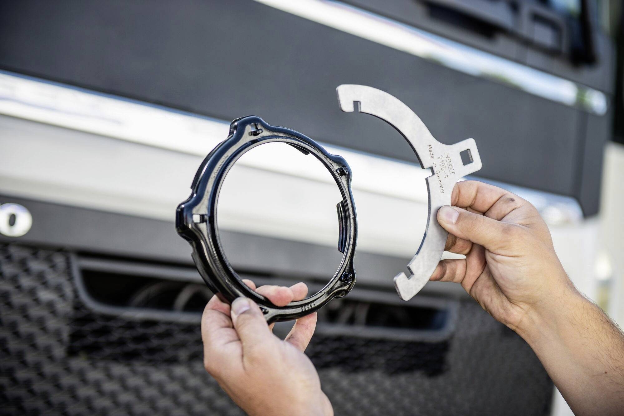 Dvě ruce drží černou prstencovou náhradní součástku a šedou konzoli před mřížkou chladiče automobilu.