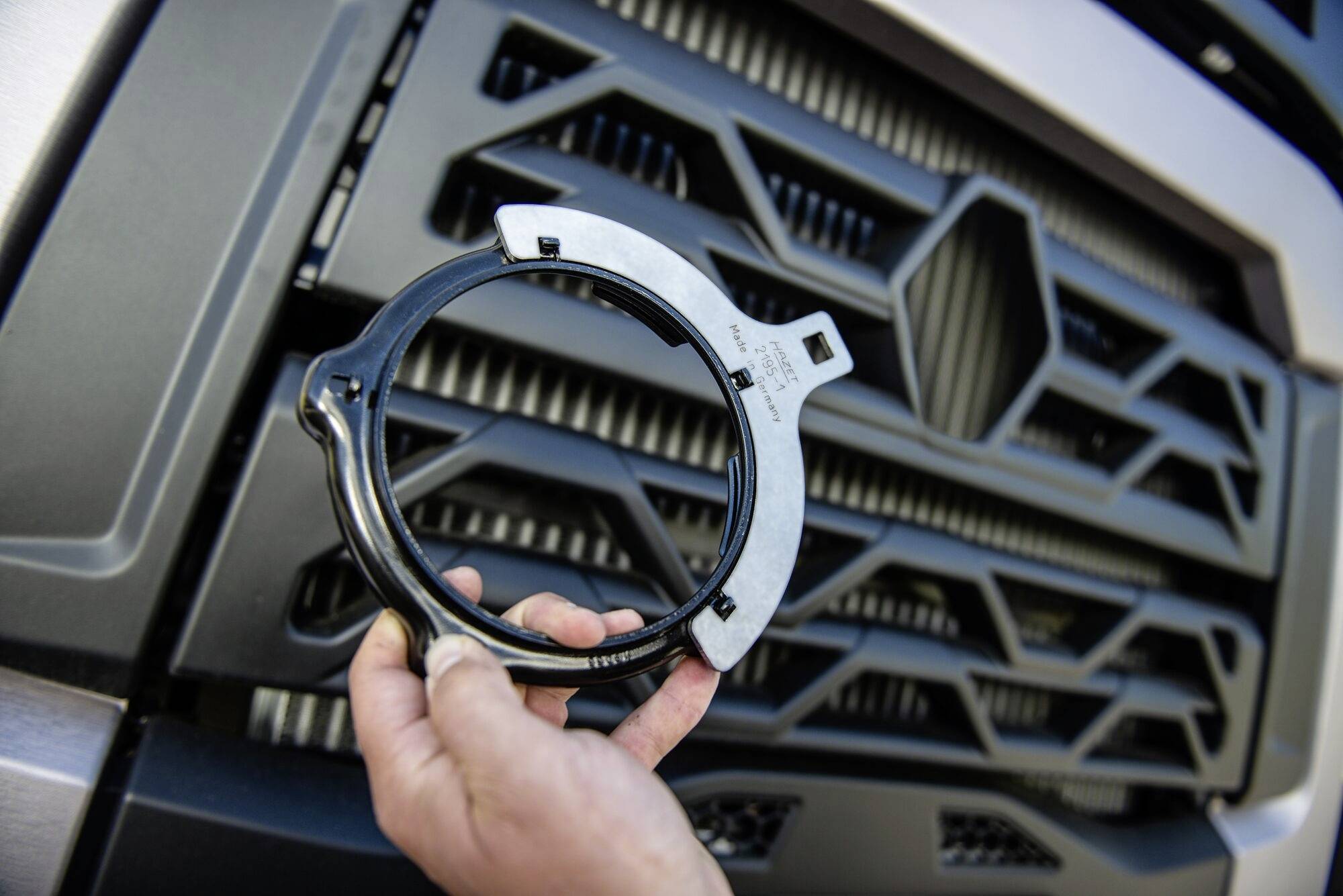 Jedna ruka drží kovový kroužek se zámkem před chladičovou mříží nákladního vozu. Kroužek vypadá jako mechanická součástka.