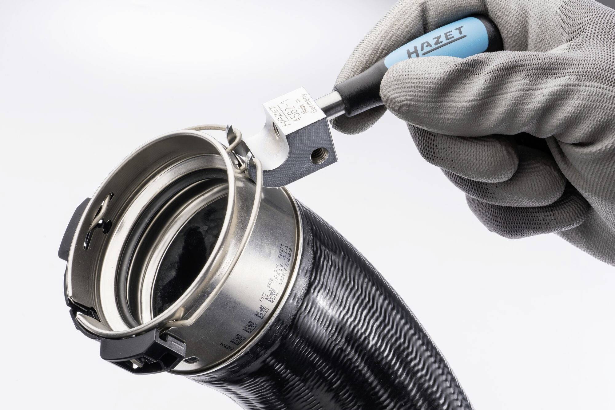 Ruka v rukavici drží modro-černý nástroj značky 'HAZET' u kovové hadicové svorky, která obepíná část potrubí.