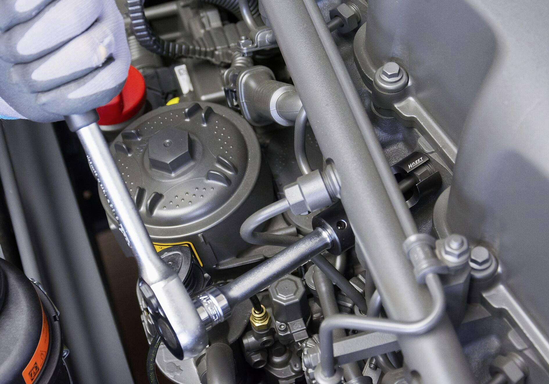 Detailní záběr mechanika, který používá ráčnu při práci na motorovém bloku, obklopený různými součástkami motoru.