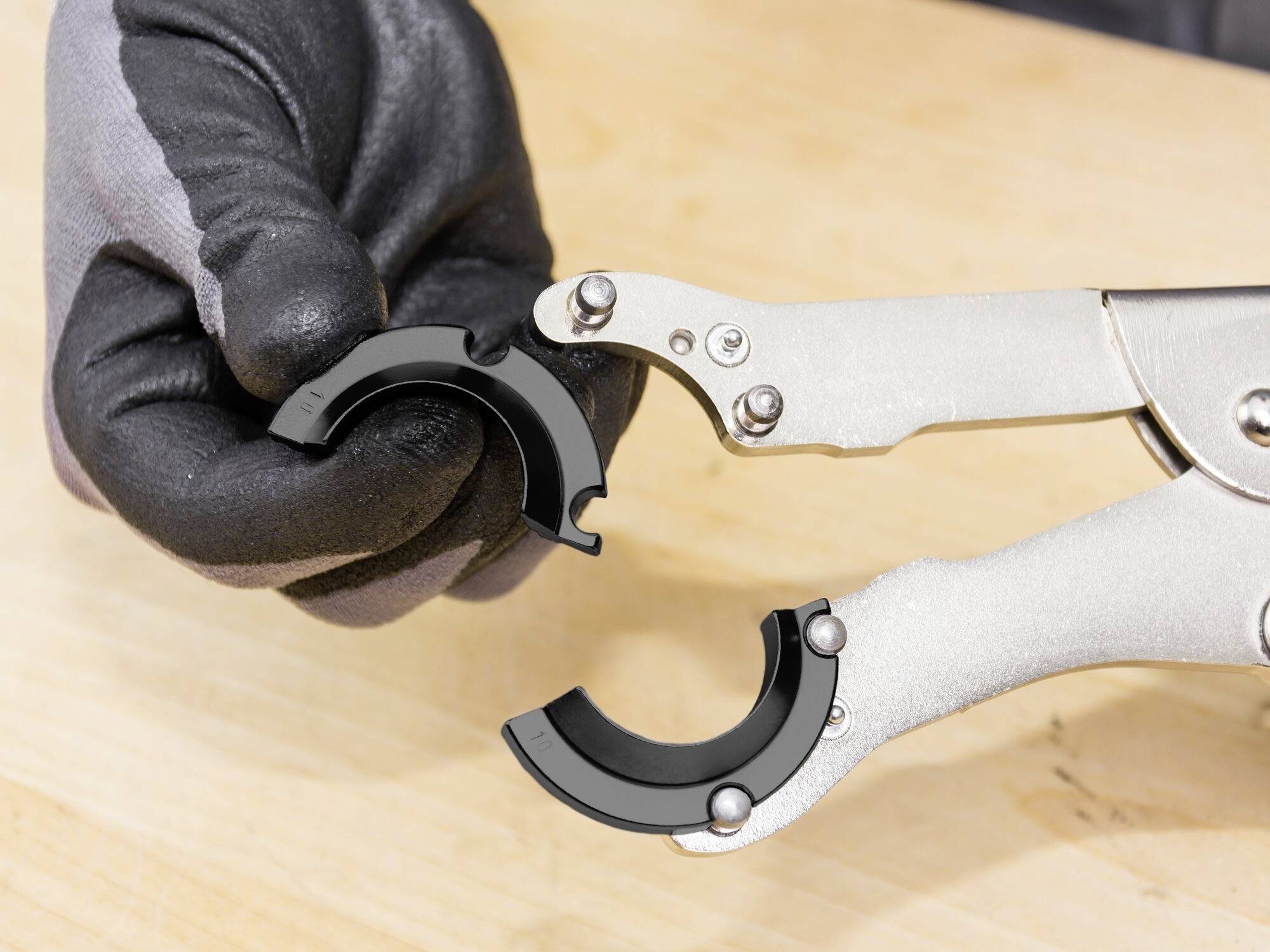 Rukavicí opatěná ruka drží kleštěmi dva půlkruhové kovové díly nad dřevěným povrchem.