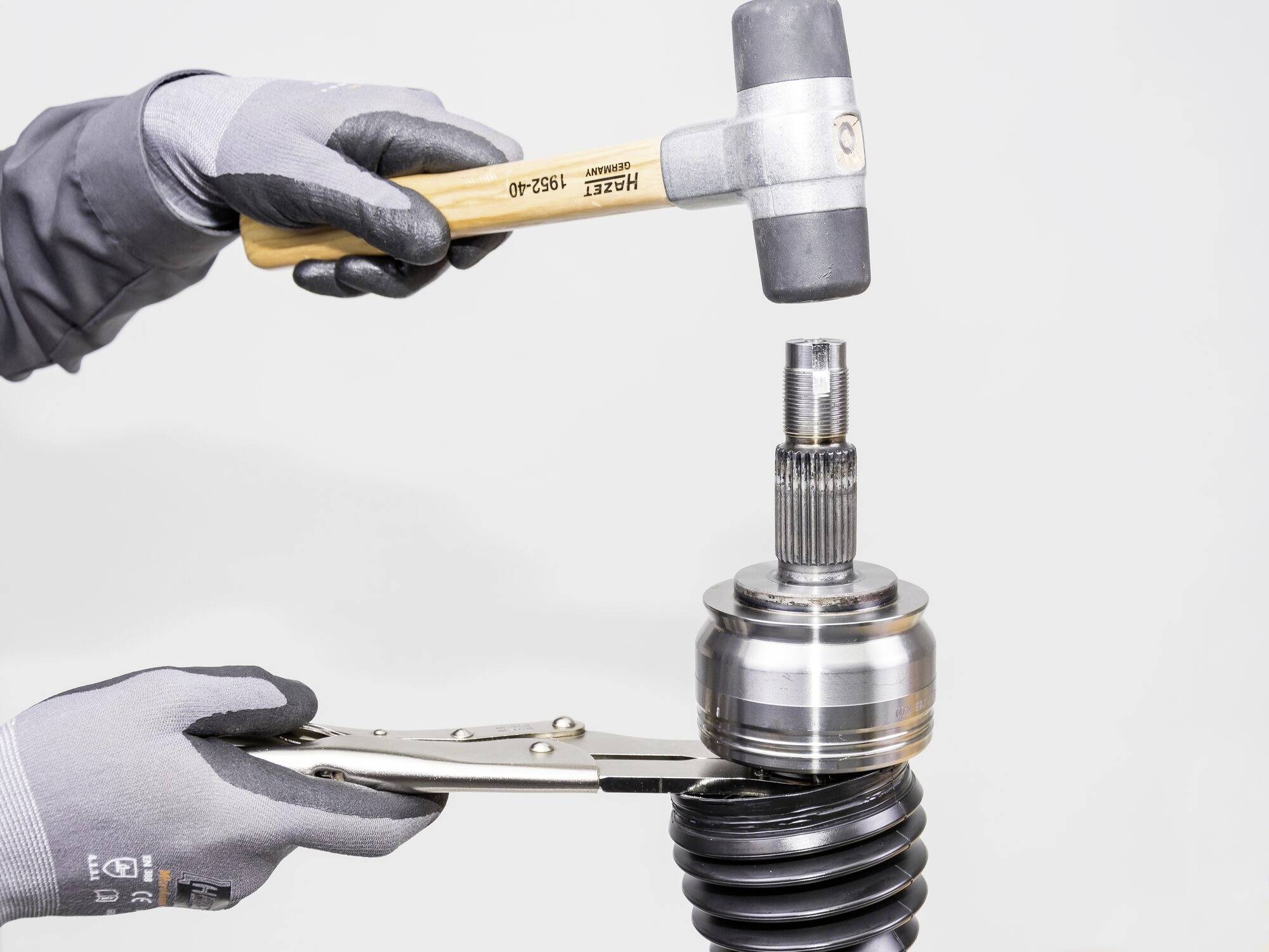 Dvě ruce v pracovních rukavicích drží nářadí: jedna svírá kladivo, druhá kleště, pravděpodobně za účelem opravy součástky, možná hnací hřídele.