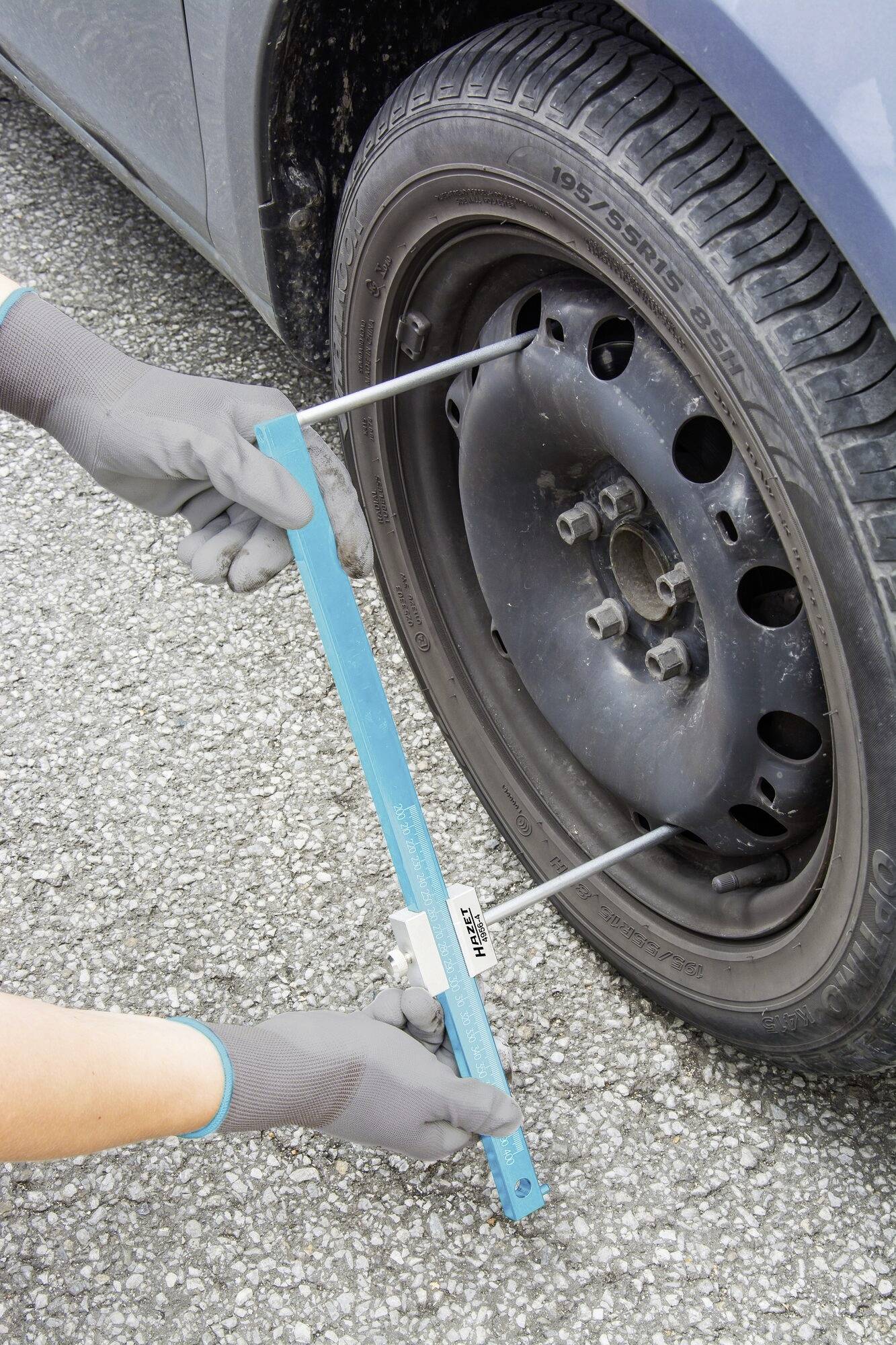 Osoba měří rozteč šroubů automobilového kola pomocí nástroje na asfaltovém povrchu.