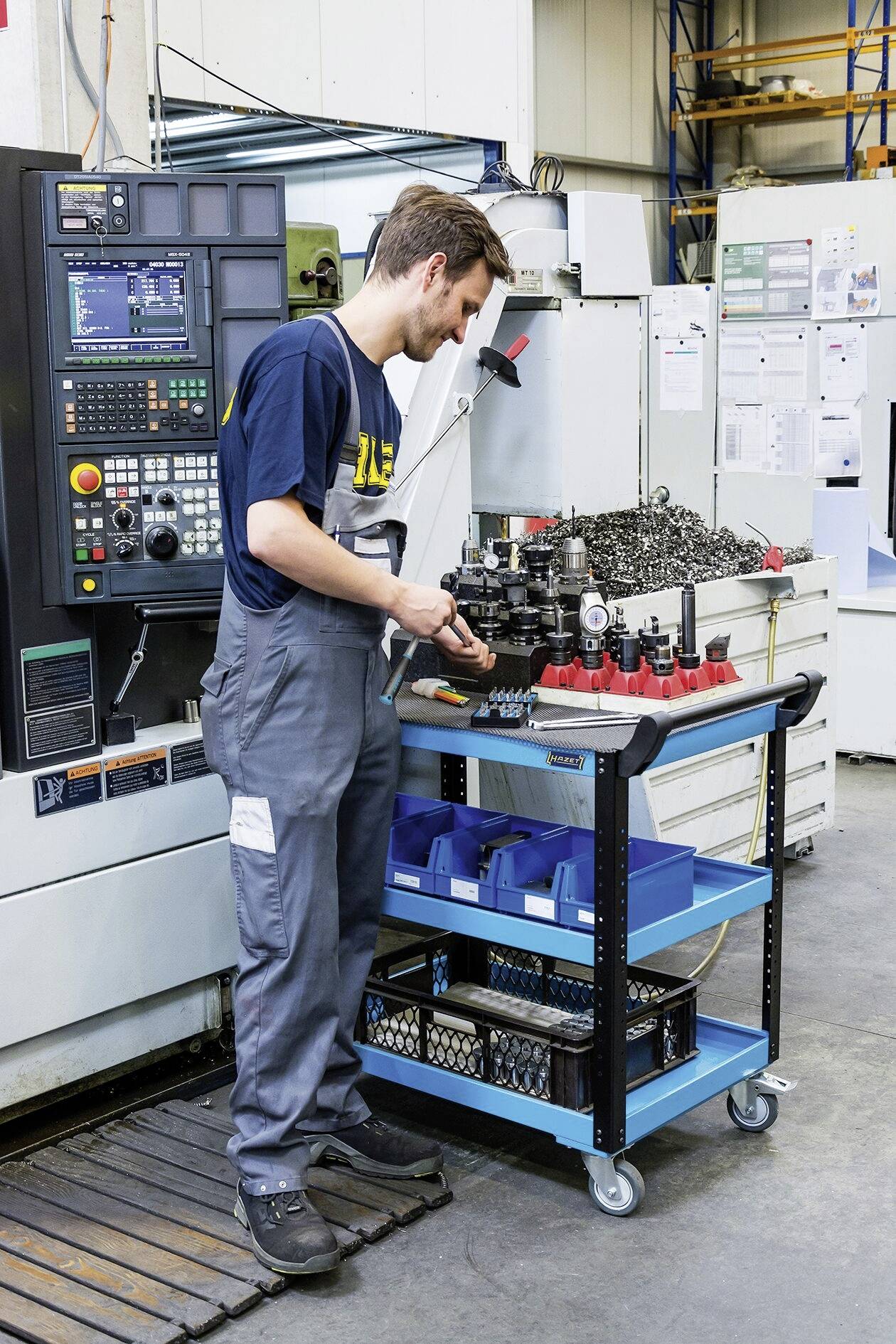 Dělník obsluhuje CNC stroj v dílně. Organizuje nástroje na vozíku poblíž stroje.