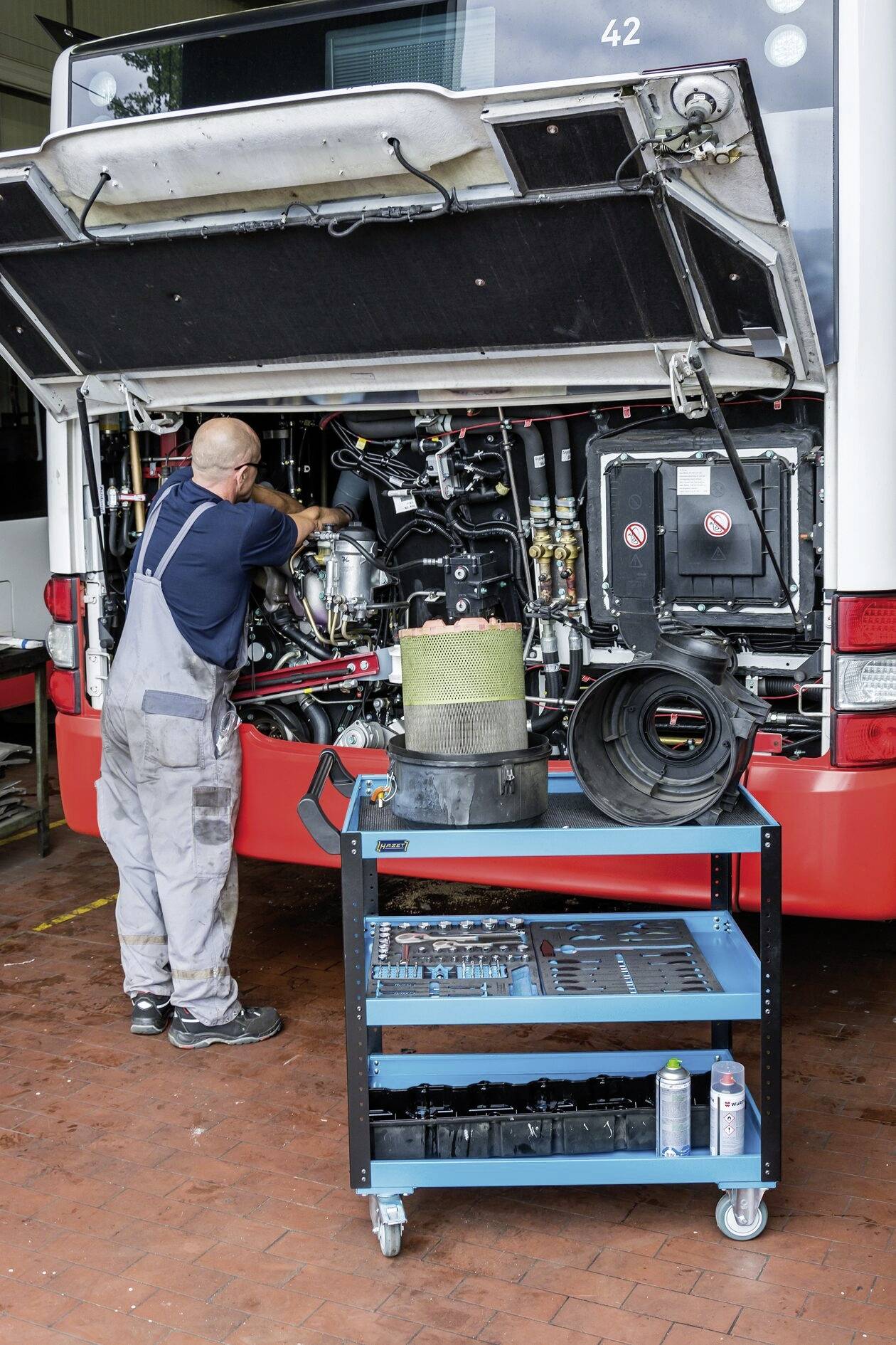 Mechanik pracuje na motoru autobusu v dílně. V popředí stojí vozík s nástroji a díly.