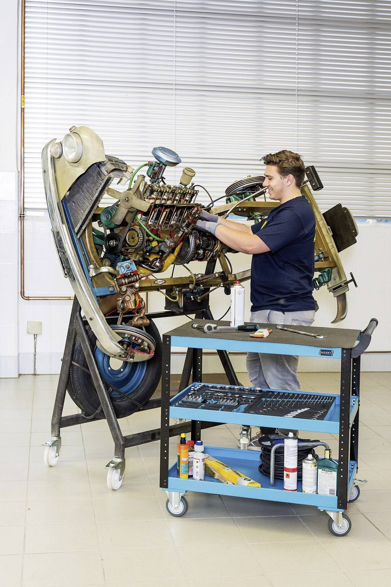 Technik pracuje na namontovaném leteckém motoru v dílně. Vedle něj stojí modrý vozík s nářadím.
