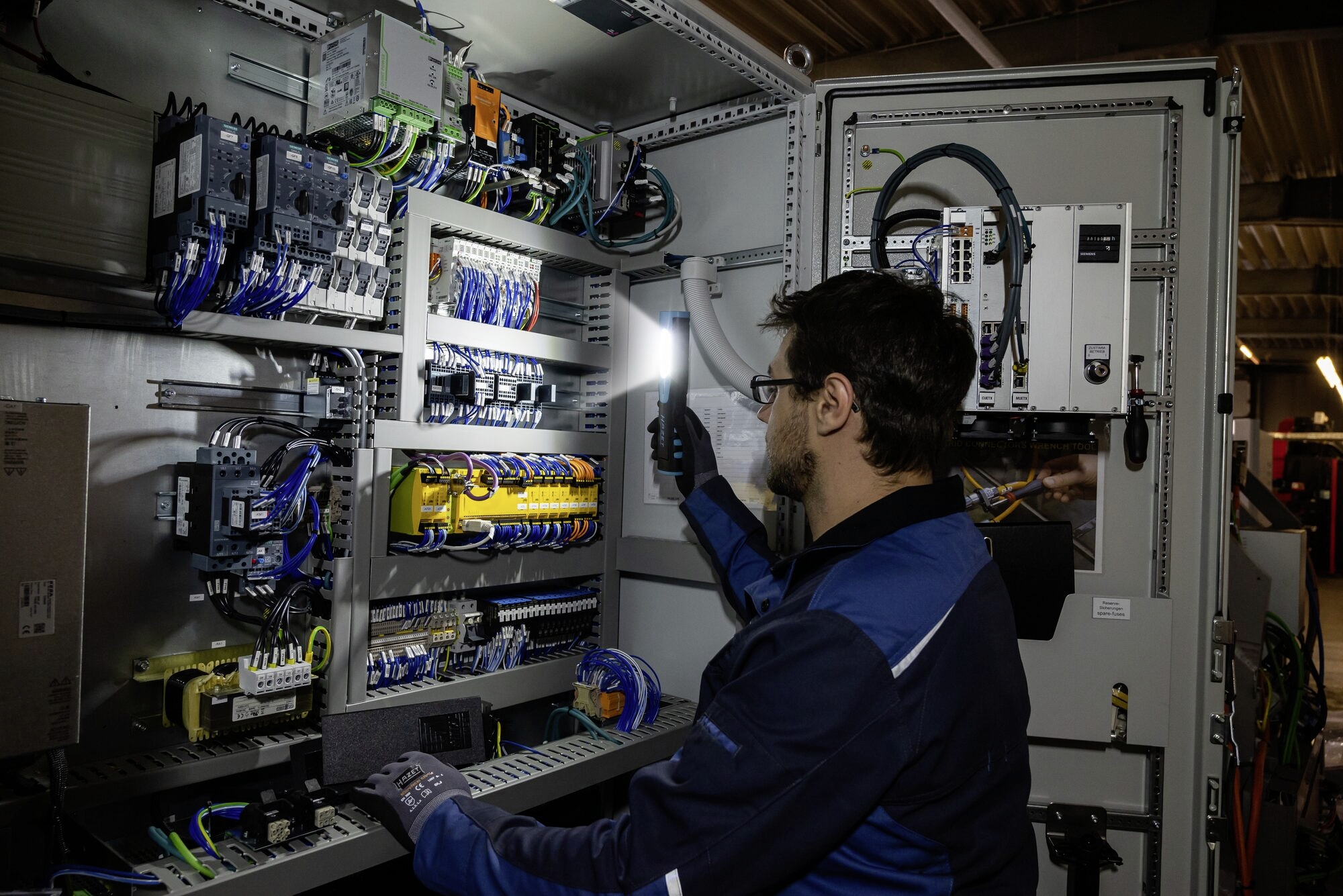 Technik pracuje s baterkou na rozvaděči, zkoumá zapojení a komponenty v průmyslovém prostředí.