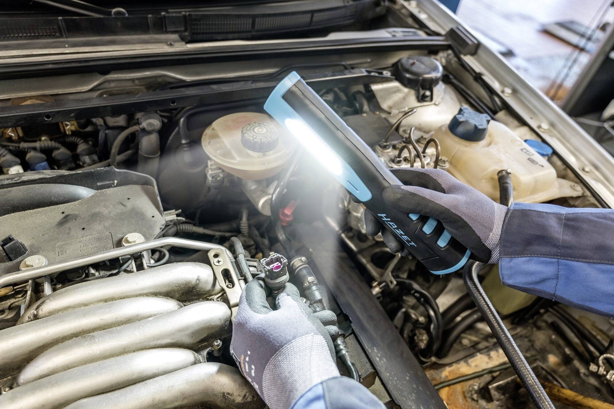 Mechanik kontroluje automobilový motor v dílně. Používá lampu, aby osvětlil motorový prostor.