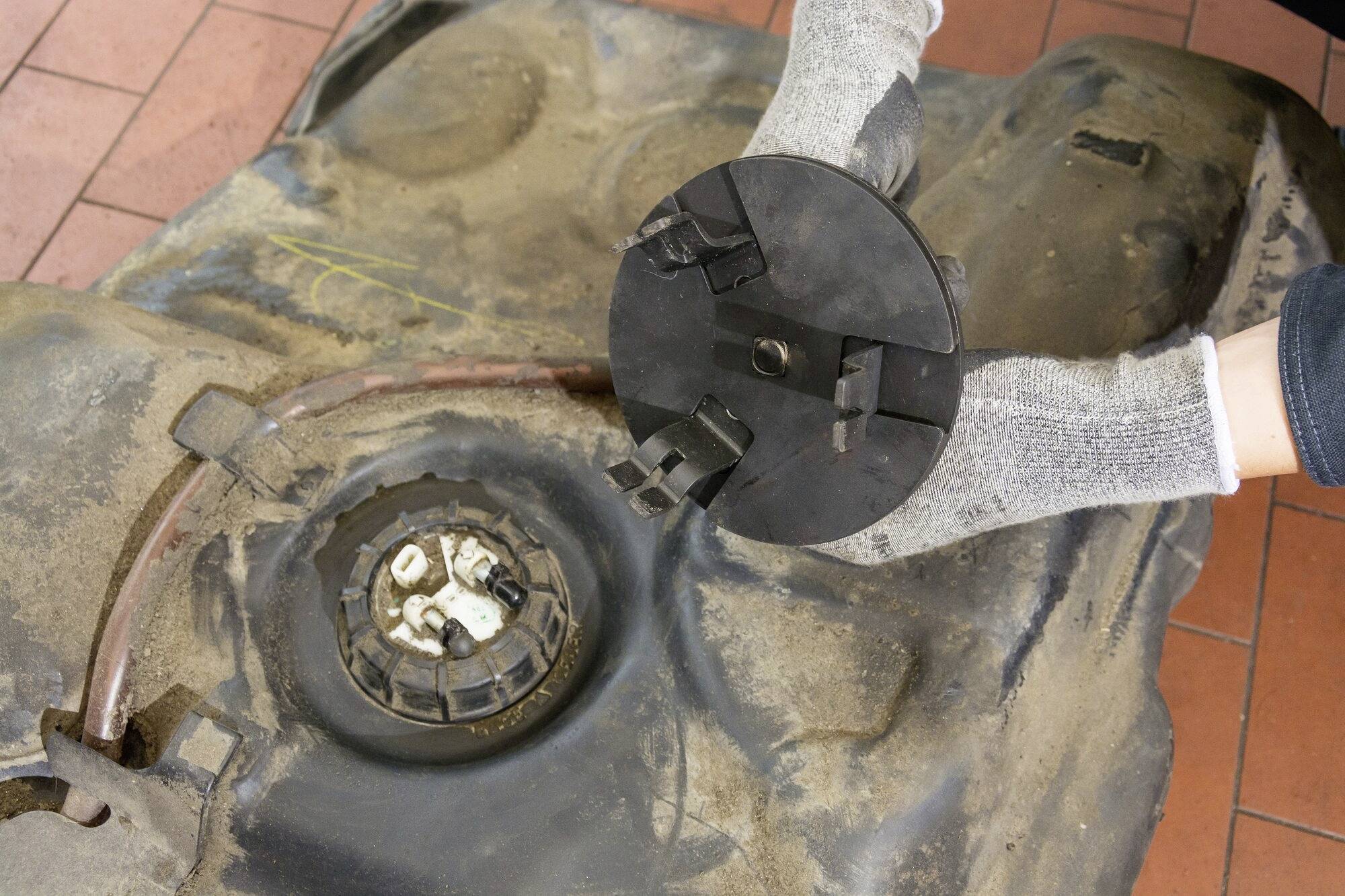 Osoba opravuje palivové čerpadlo v automobilu. Pravá ruka drží víko čerpadla, zatímco levá ruka pracuje na znečištěné nádrži.
