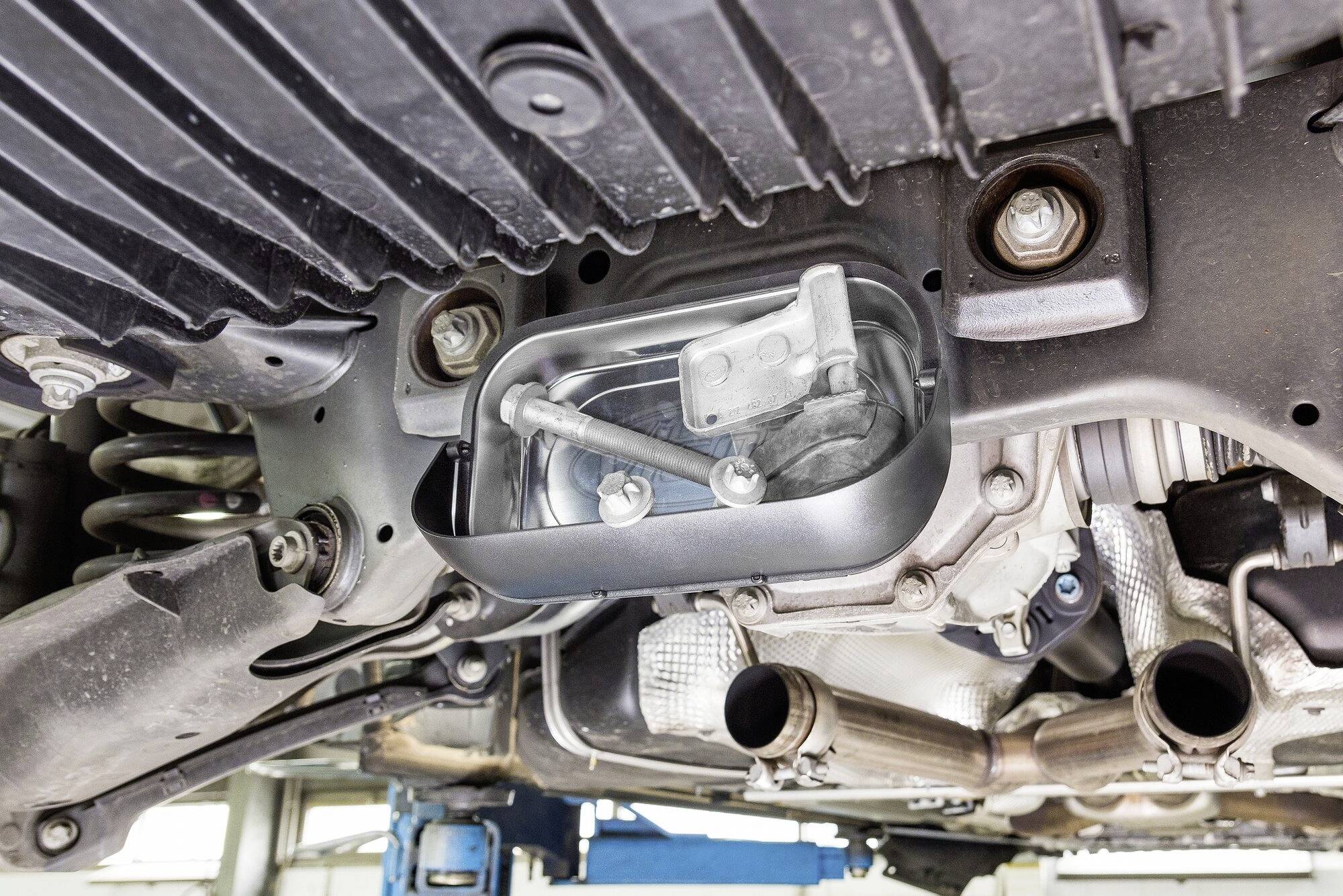 Spodní strana vozidla s odhaleným motorem a viditelnými komponenty v prostředí autoservisu.