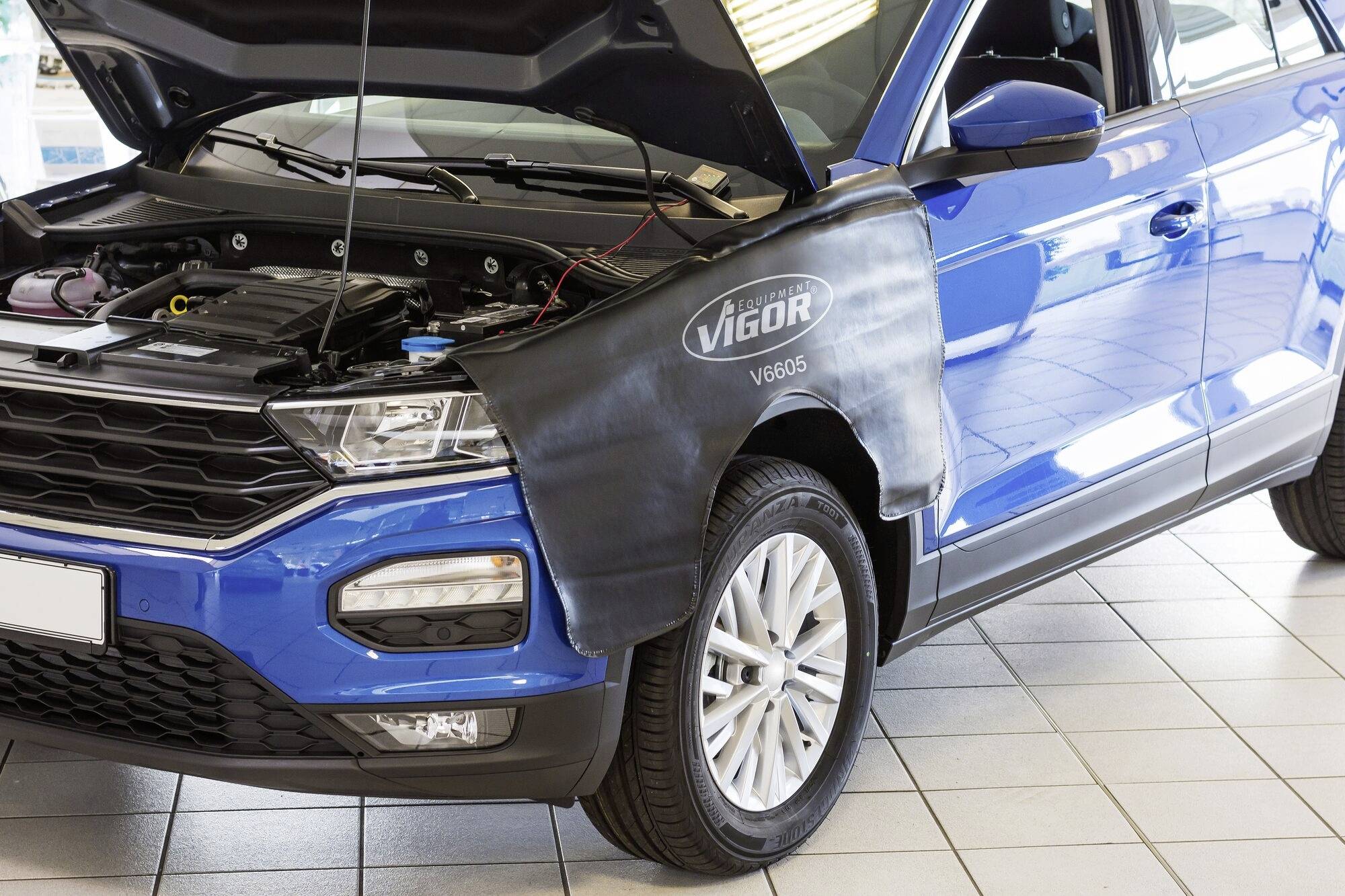 Modrý osobní automobil se zvednutou kapotou stojí v autoservisu. Na blatníku je umístěn černý ochranný kryt s nápisem 'VIGOR'.