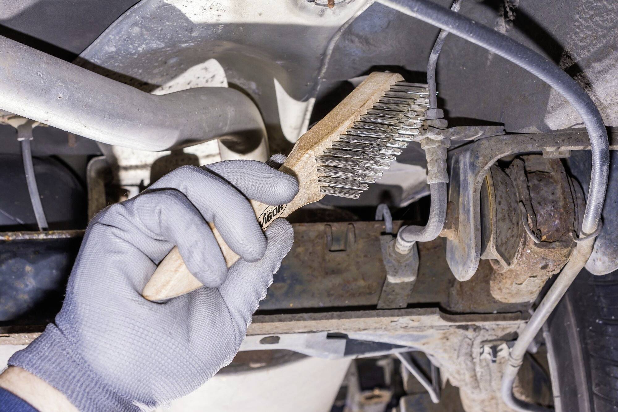 Ruka v rukavici čistí spodek automobilu drátěným kartáčem se zaměřením na odstraňování rzi.