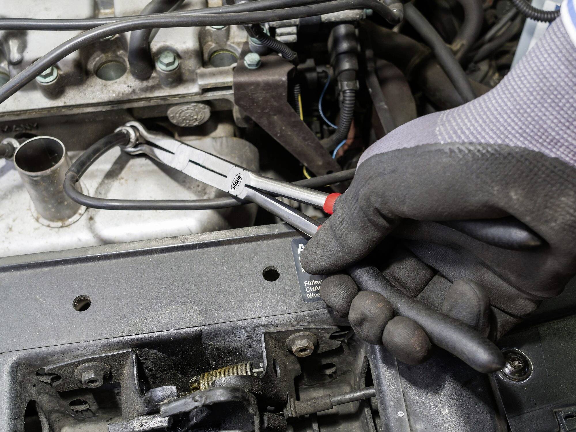 'Detailní záběr mechanika, který zasahuje kleštěmi do automobilové součásti. V pozadí je viditelný motorový prostor.'