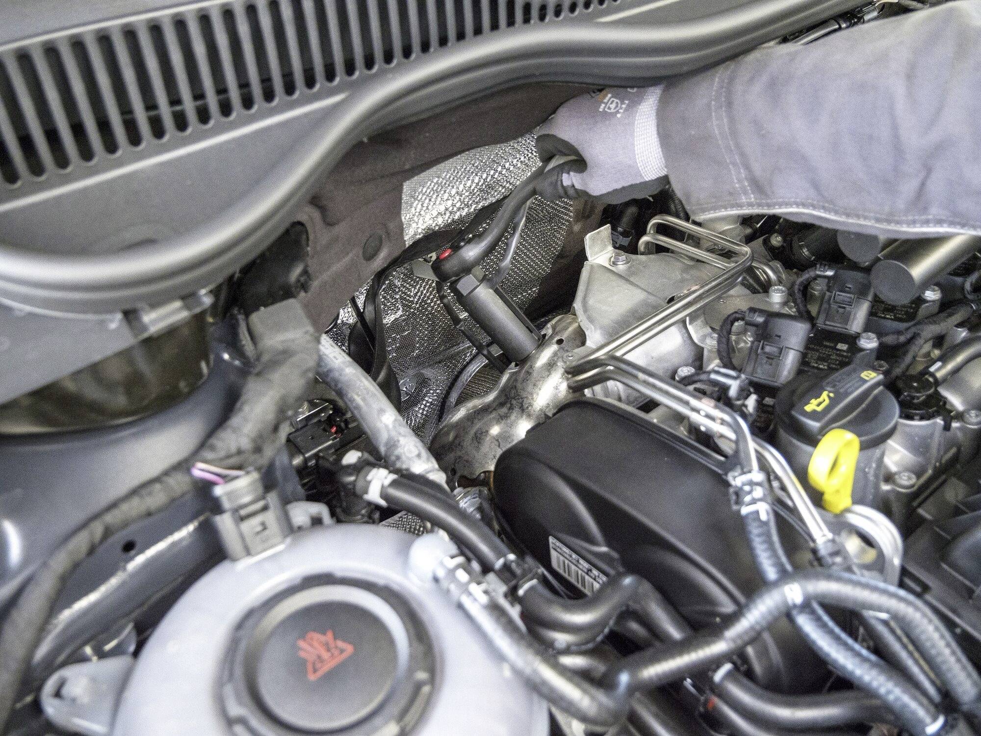 Osoba kontroluje motorové spoje v automobilu. Pod kapotou jsou viditelné různé kabely a komponenty motoru.