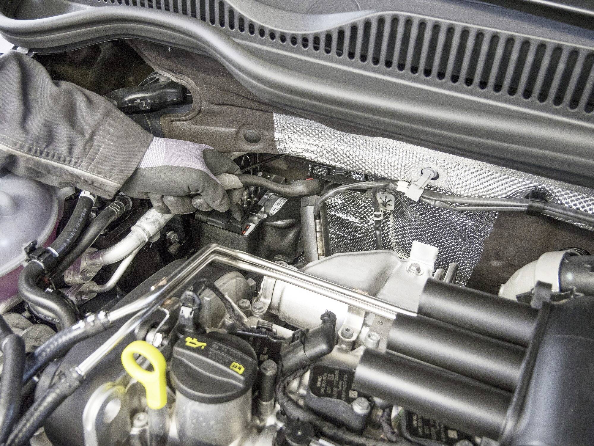 Mechanik kontroluje motorový prostor automobilu. Jsou viditelné různé hadice a komponenty. Ruka je v rukavici.