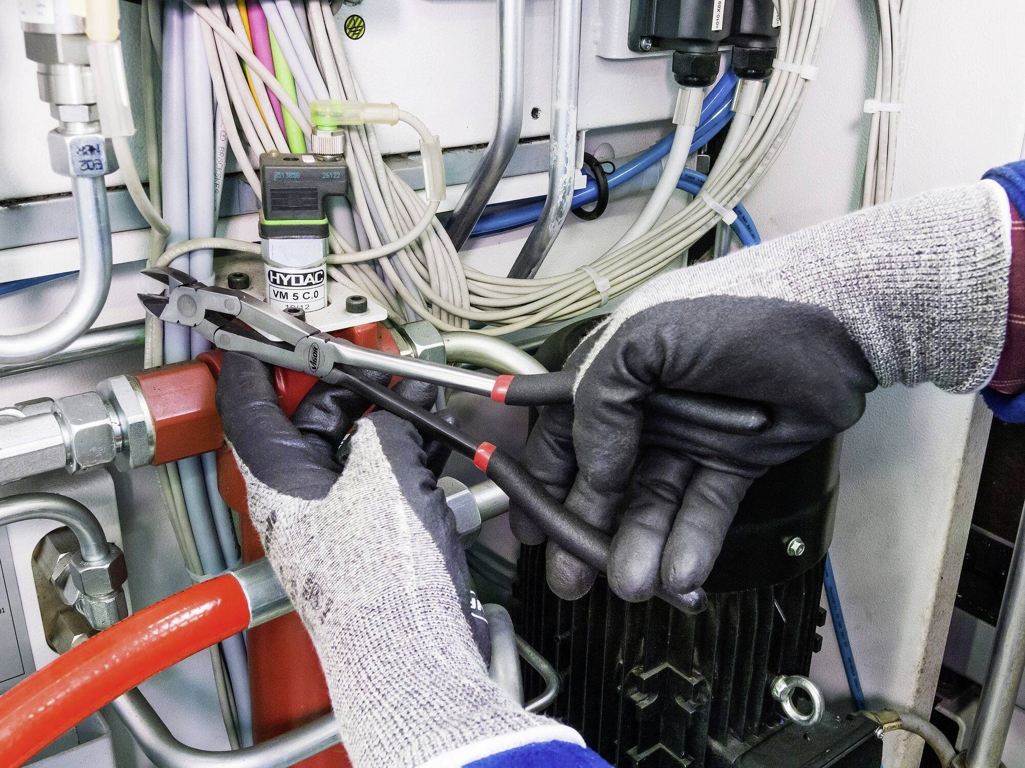 Osoba v šedých rukavicích opravuje elektrický rozvaděč. Jsou zde zobrazeny kabely a kleště.