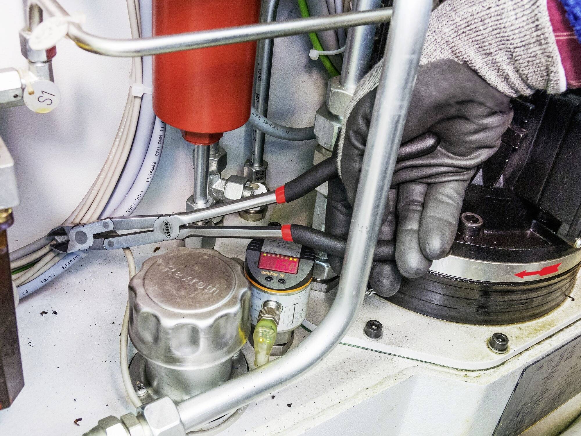 Osoba v rukavicích pracuje na hydraulickém stroji, obklopena kovovými trubkami a kabely.