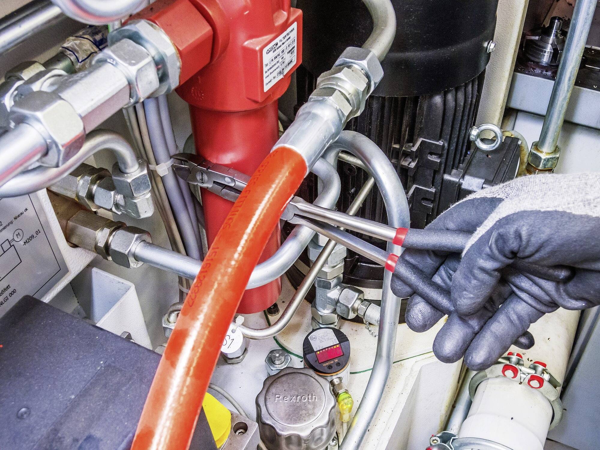 Technik utahuje šroub na hydraulickém systému pomocí klíče. Jsou viditelné červené a stříbrné trubky.