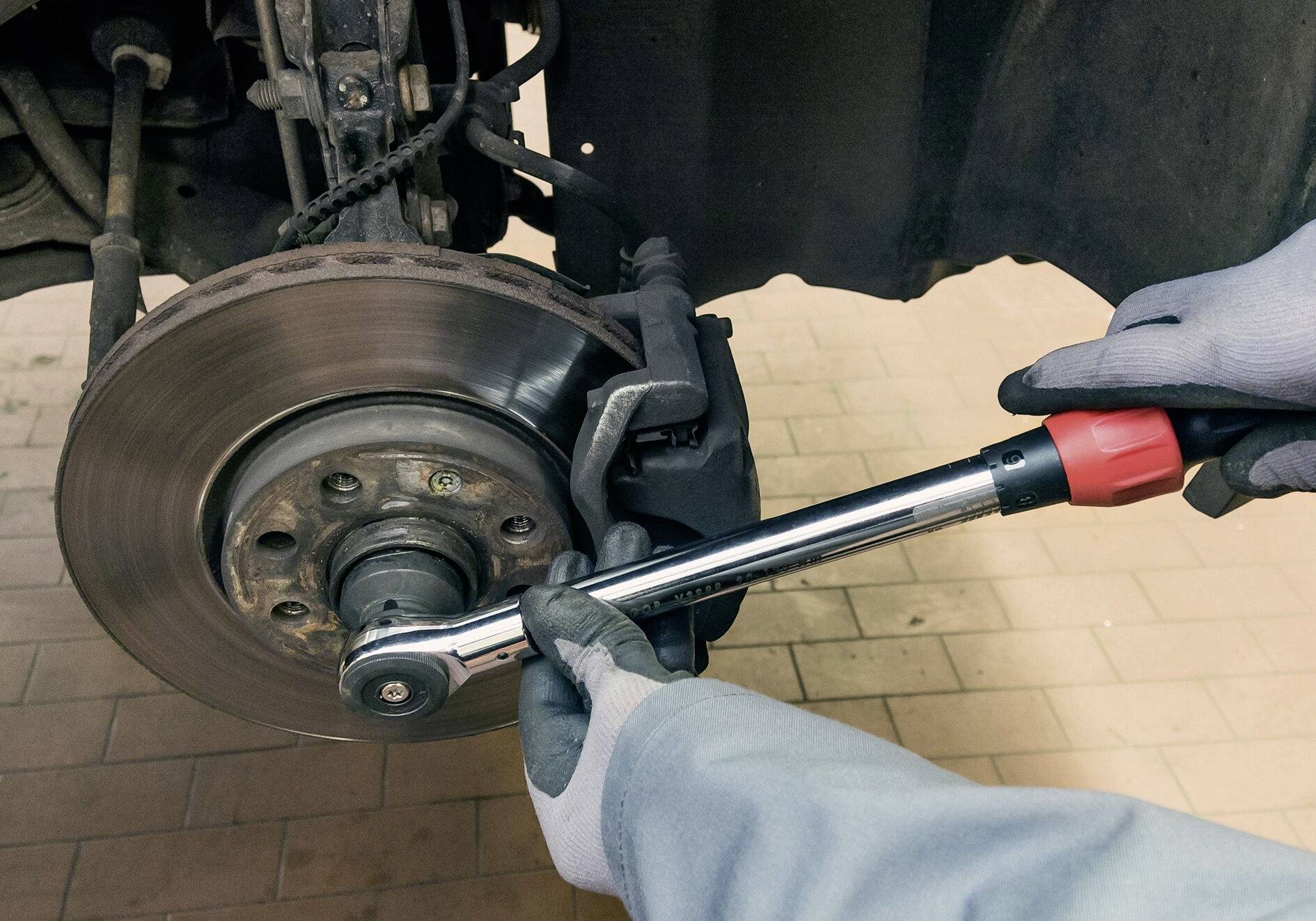 Klíč utahuje matice na brzdovém kotouči, ruce jsou v mechanických rukavicích, v pozadí je vidět interiér opravny.