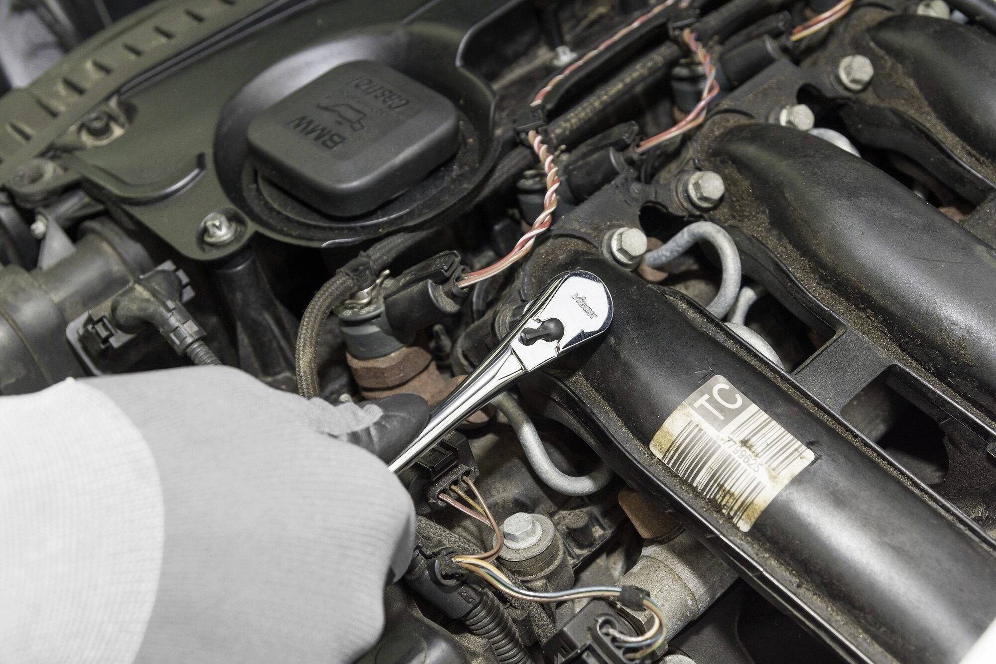 Osoba opravuje motor pomocí klíče. Důraz je kladen na motorové součásti a ruku s nástrojem.