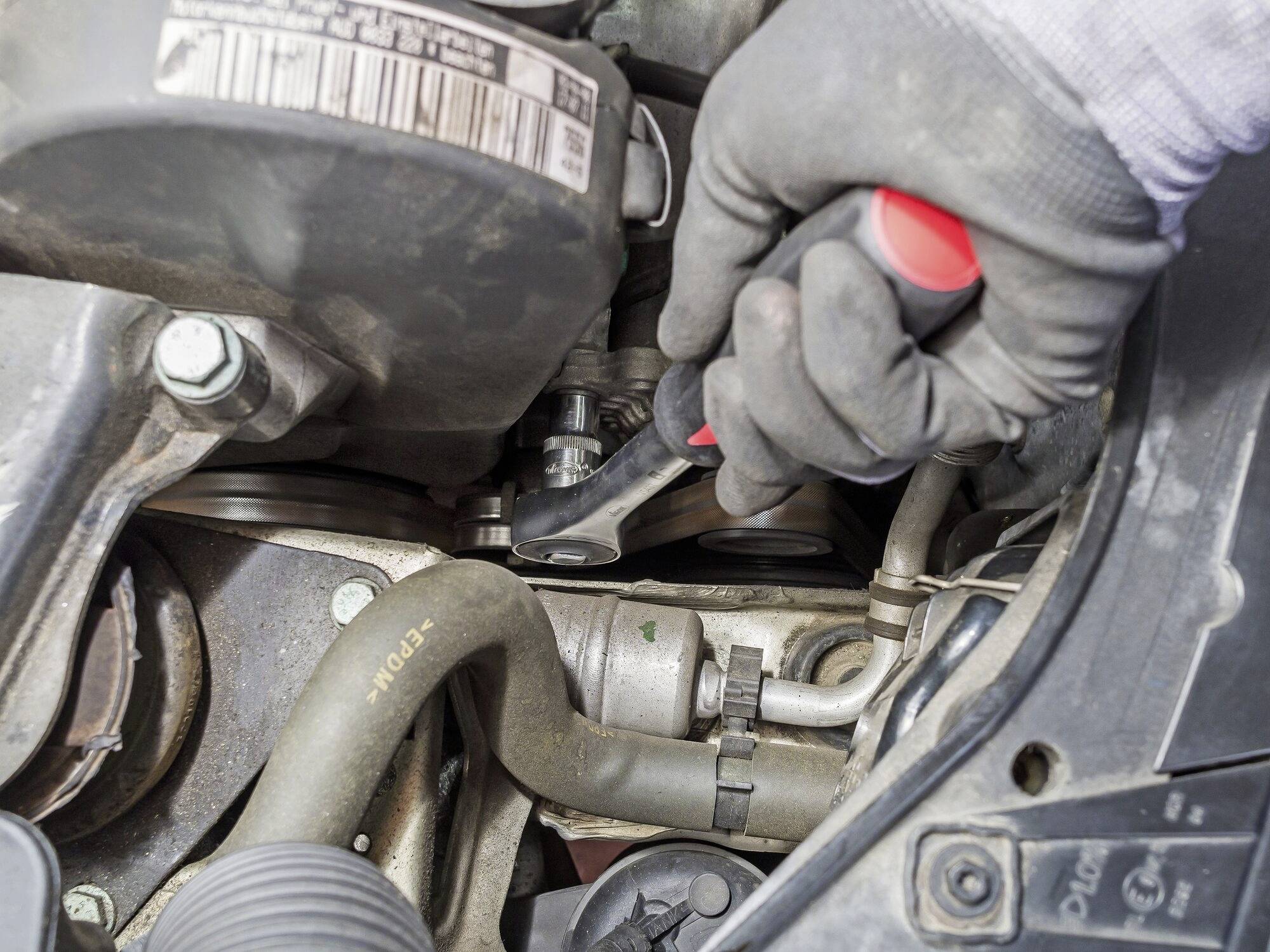 'Detailní záběr ruky v rukavici, která pracuje s klíčem na motoru vozidla. Jsou viditelné různé komponenty motoru.'