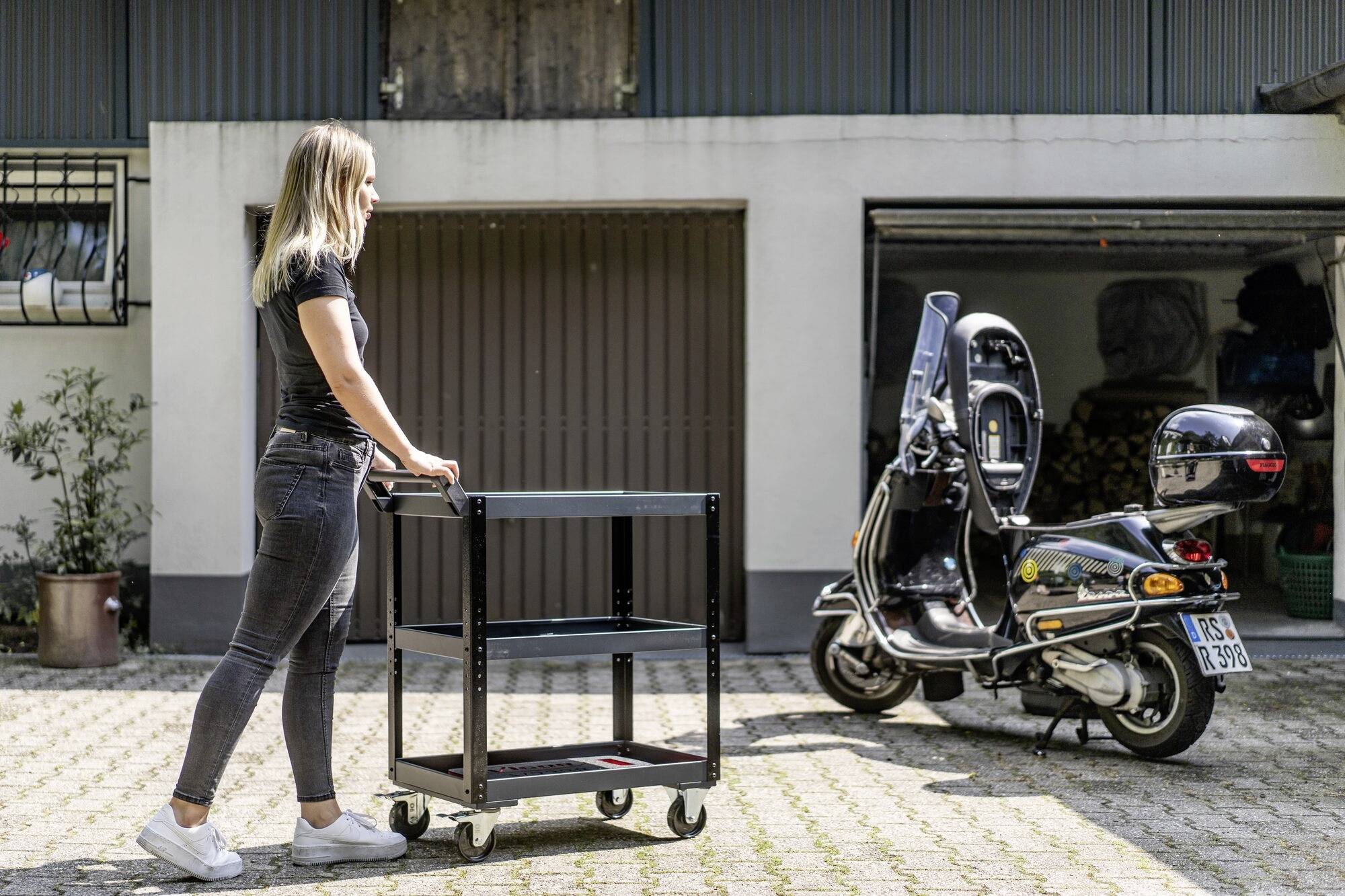Žena tlačí vozík po dlážděném dvoře před garáží. V pozadí stojí skútr.