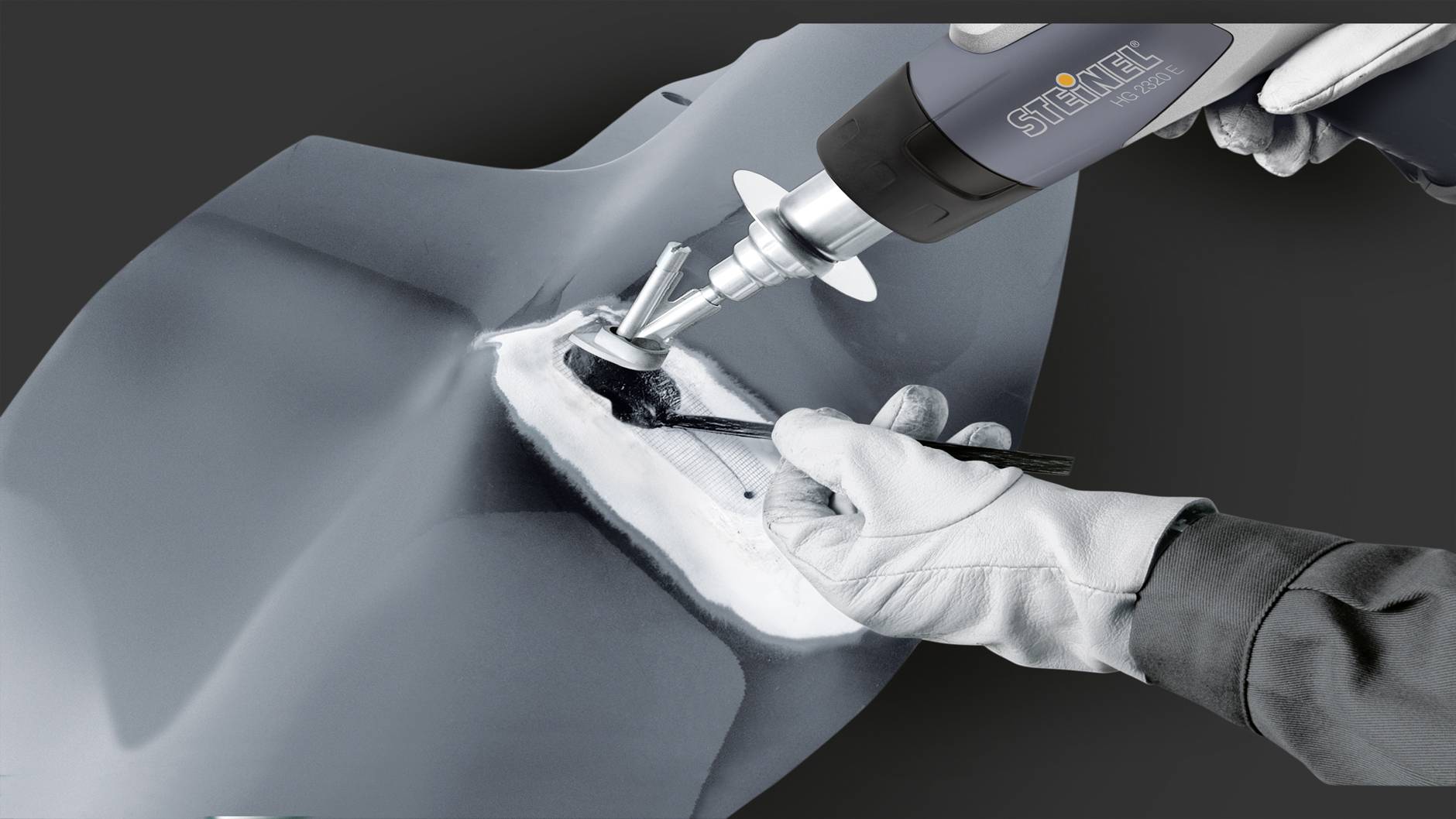 Osoba opravuje poškozenou část kovového dílu pomocí horkovzdušné pistole. Bezpečnostní vybavení je viditelné.