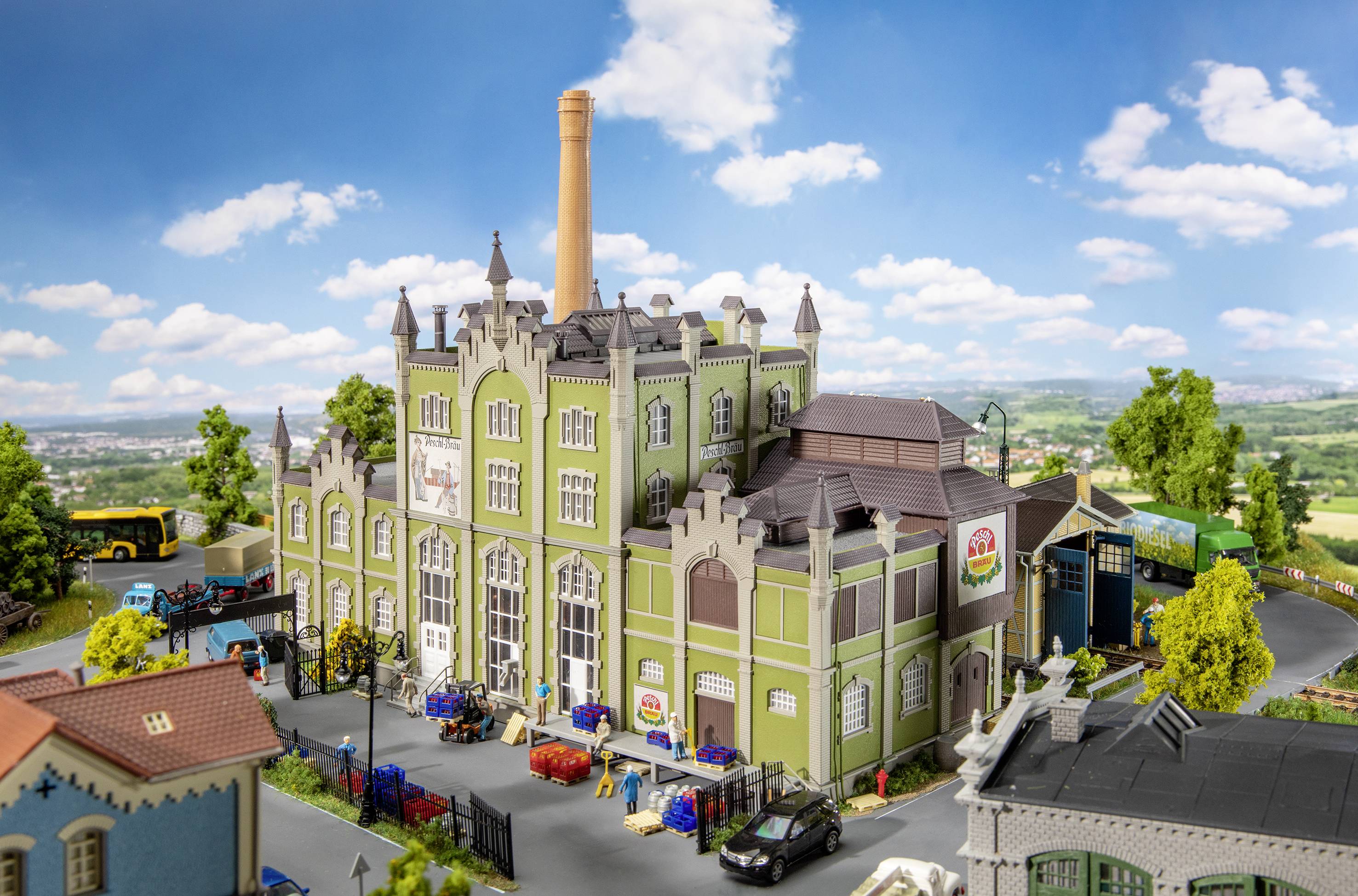 Model historické továrny se zelenými fasádami, obklopený miniaturními vozidly a stromy, pod modrým nebem.