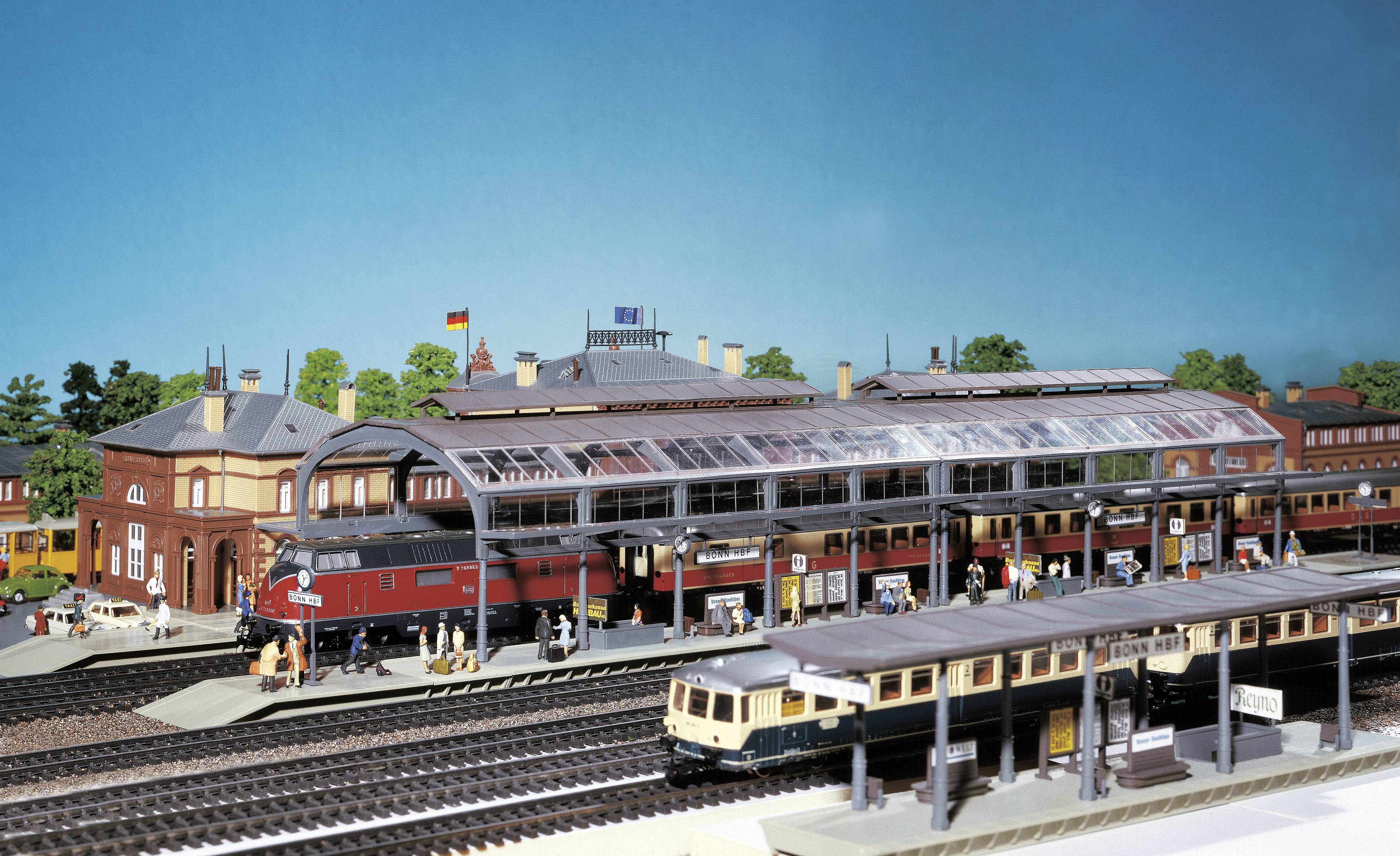 Modelové kolejiště s lokomotivami, vlaky a figurkami na nástupištích. Detailní zobrazení nádraží s historickými vlaky.