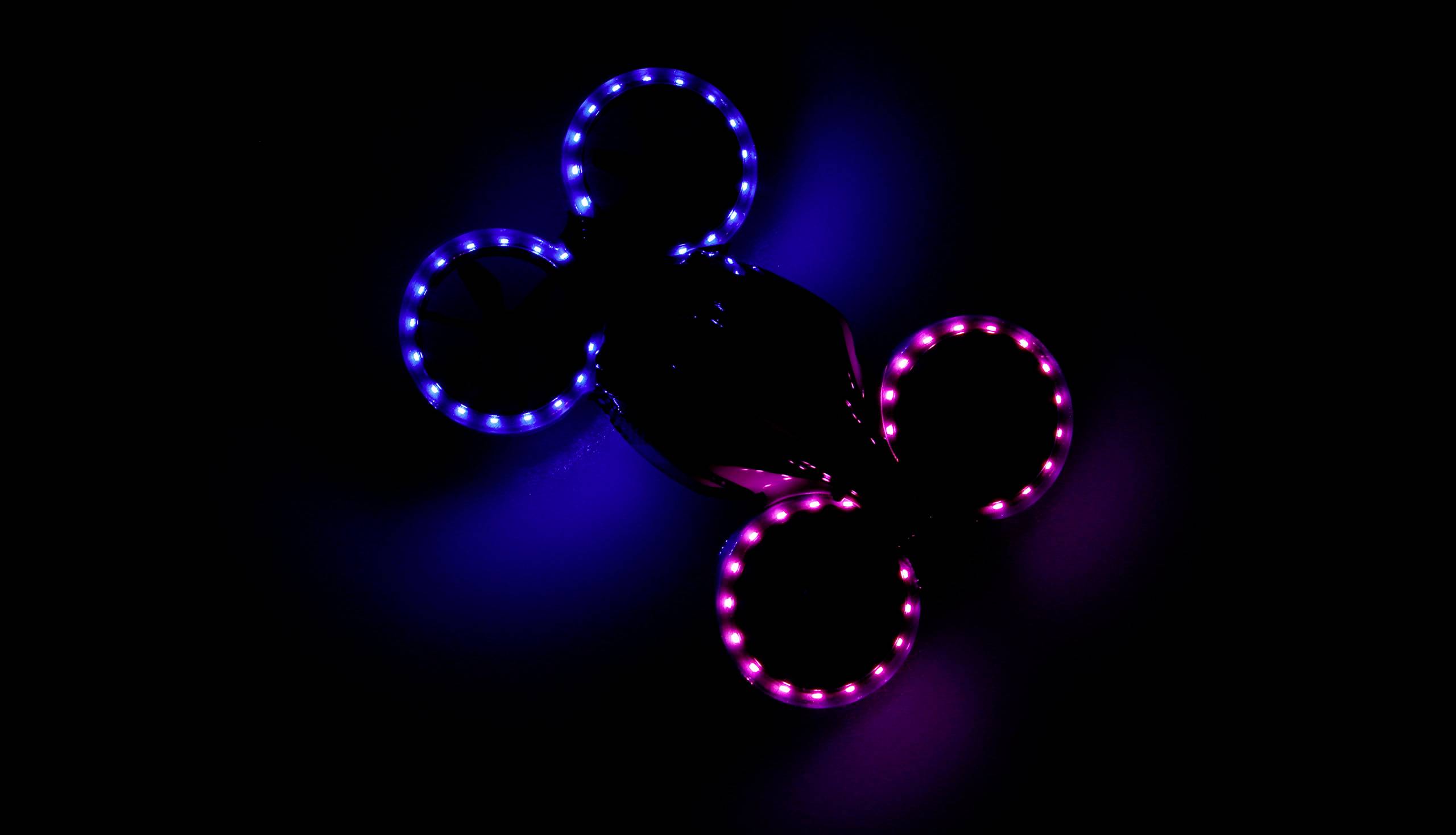 Dron se čtyřmi svítícími rotory, dva v modré a dva v purpurové barvě, letí v tmavém prostředí.