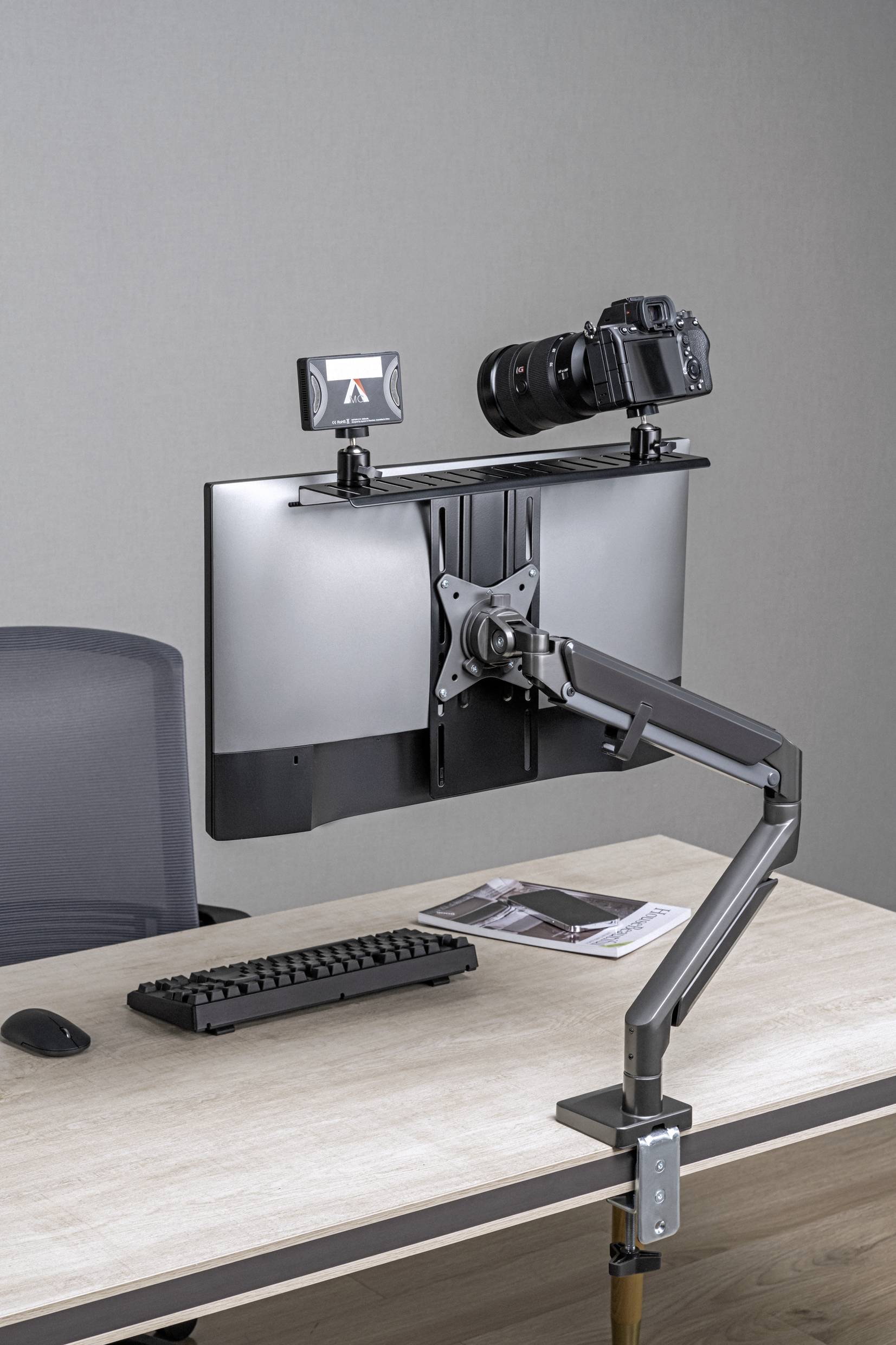 Počítačový monitor na nastavitelném rameni umístěný na psacím stole, s připevněnou kamerou a malým monitorem.