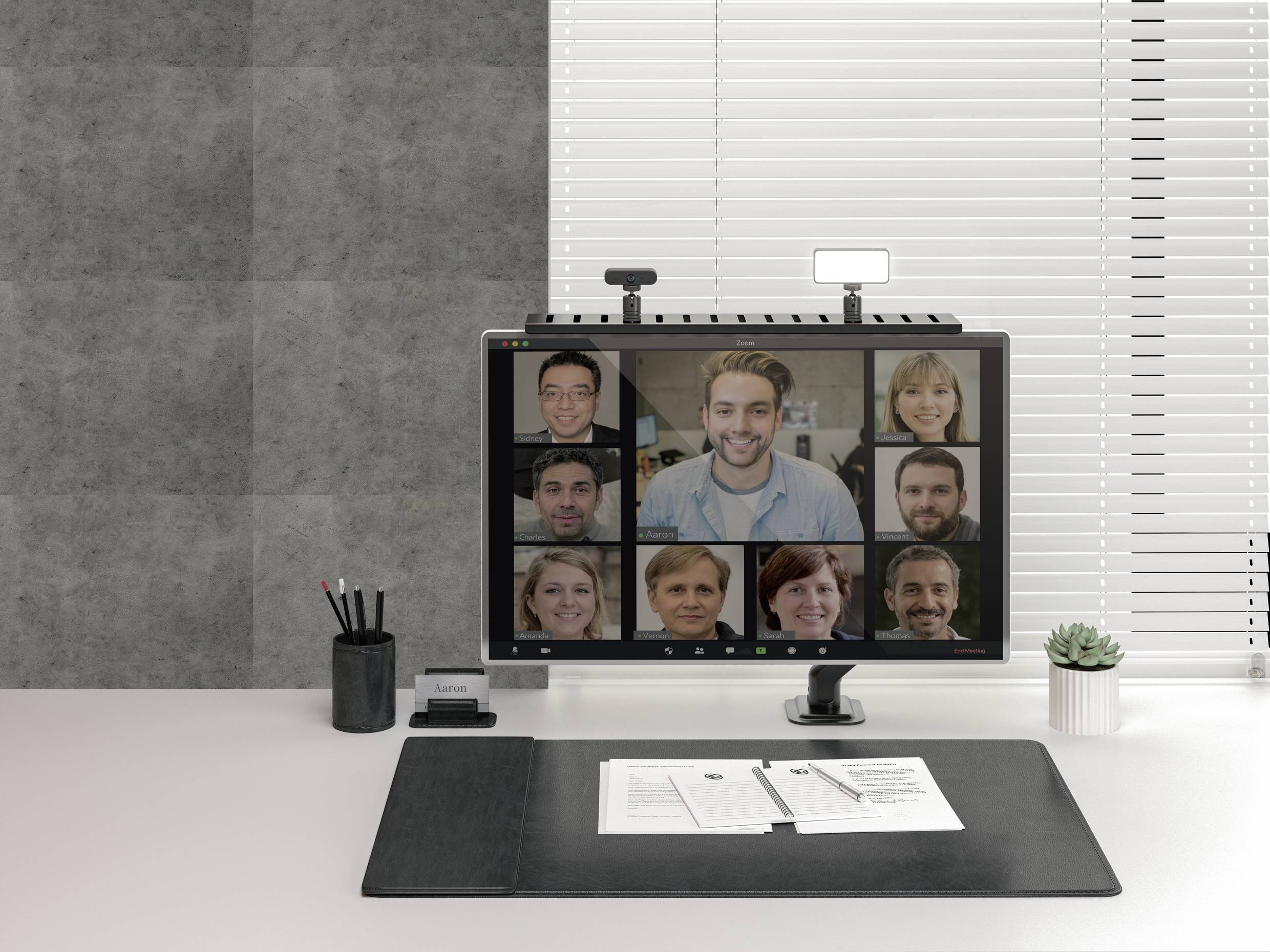 Počítačový monitor zobrazuje videokonferenci s devíti účastníky. Místnost je jasně osvětlená, na stole jsou položeny tužky a poznámkový blok.