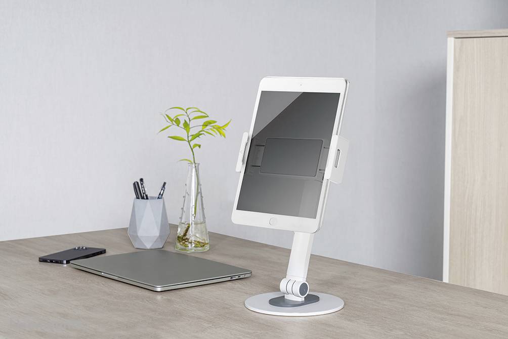 Držák tabletu na minimalistickém pracovním stole s laptopem, smartphonem, stojánkem na tužky a rostlinou.